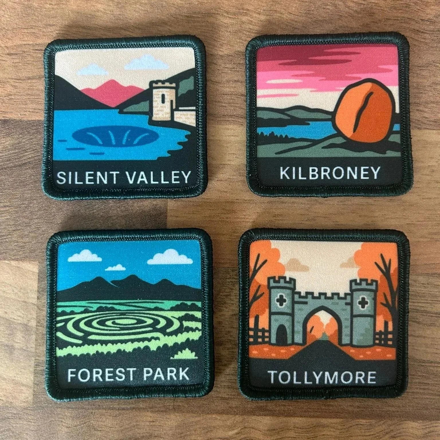 Four embroidered patches depicting scenic landscapes of Silent Valley, Kilbroney, Forest Park, and Tollymore, arranged in a 2x2 grid on a wooden surface.
