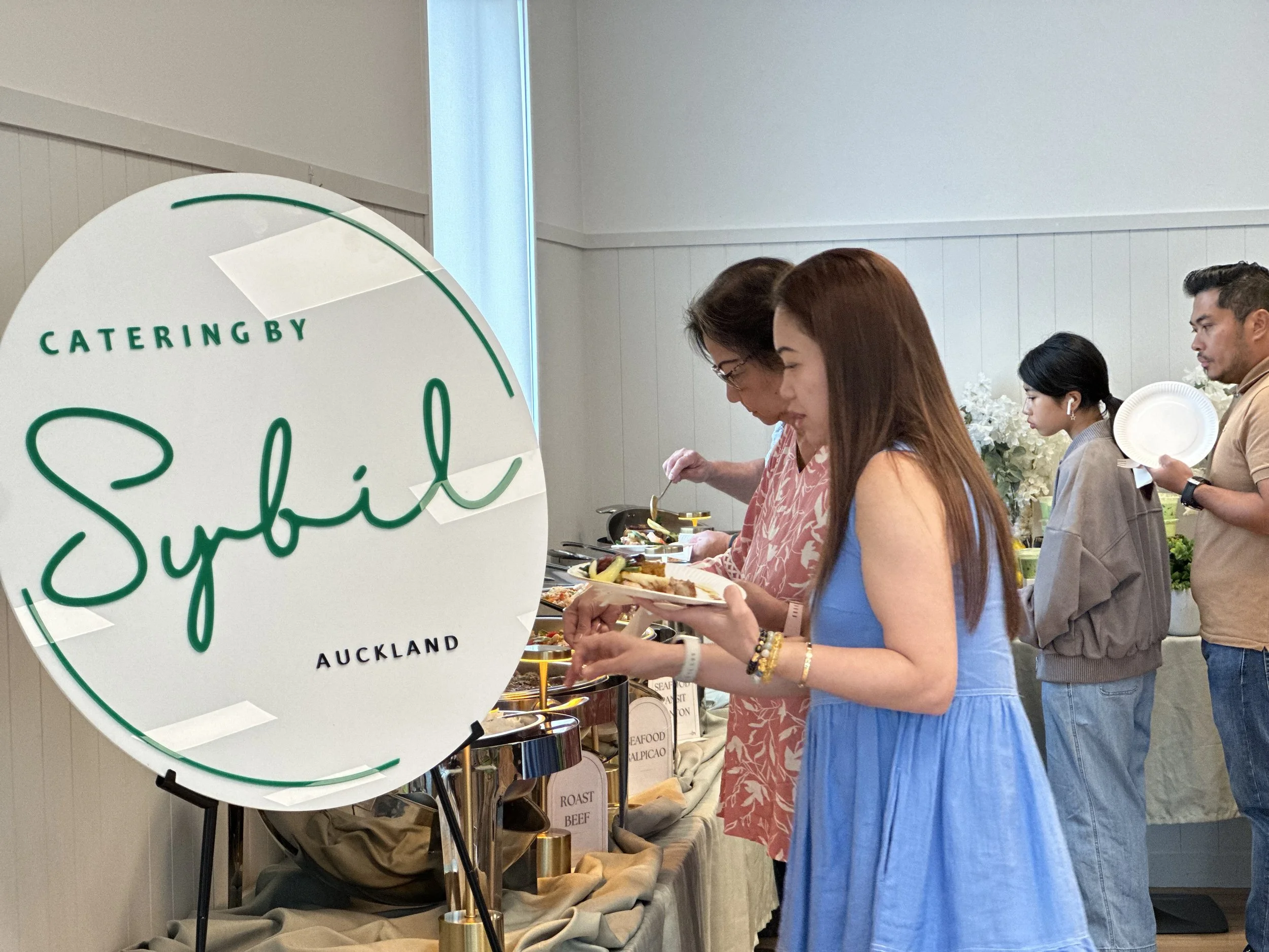 People at a buffet table serving themselves food at an event with a sign that reads "Catering by Sybil Auckland."