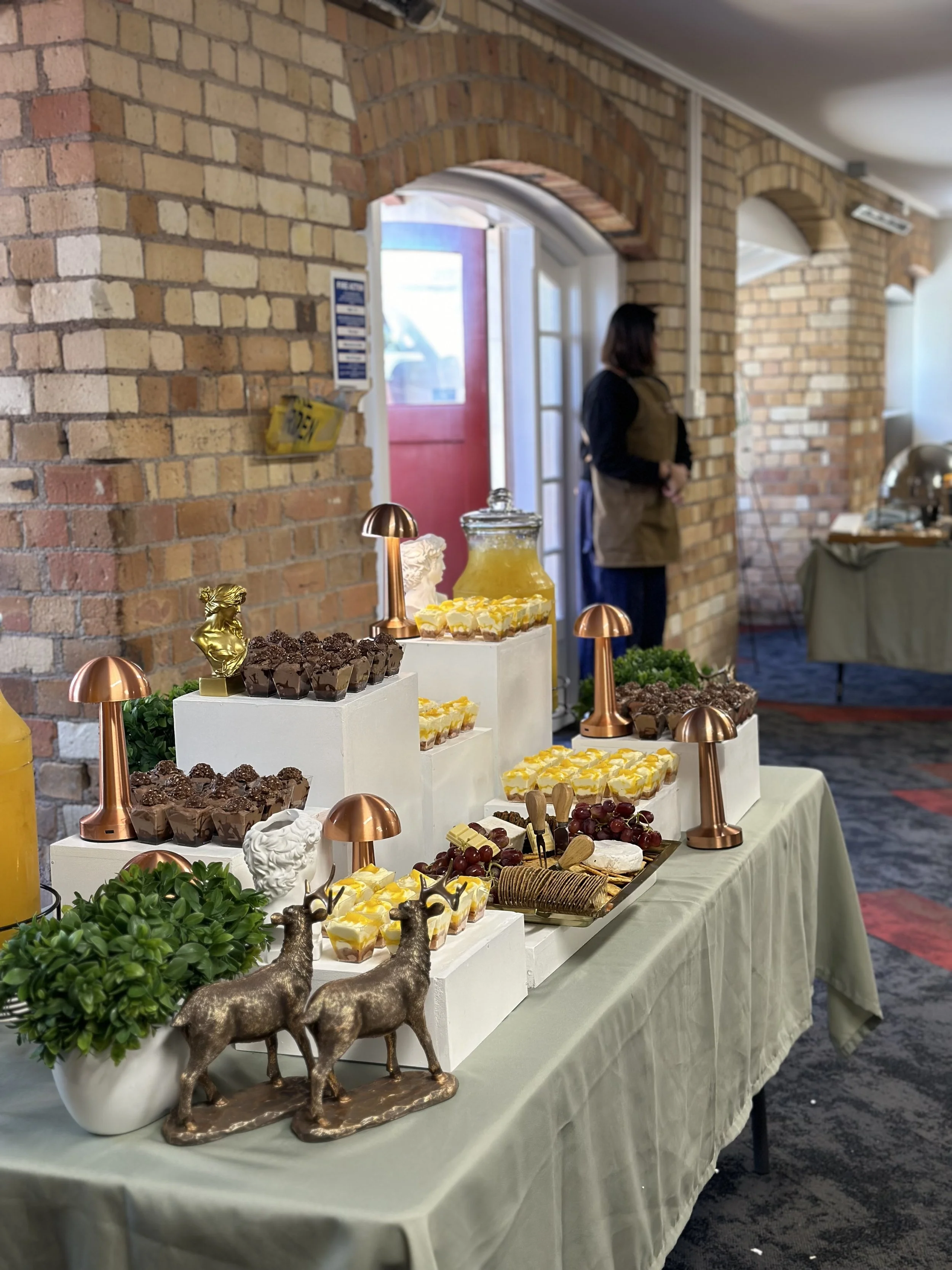 A table with decorative lamps, chocolate desserts, grapes, and a pitcher of lemonade in a brick-walled room, with a woman standing by the doorway in the background.