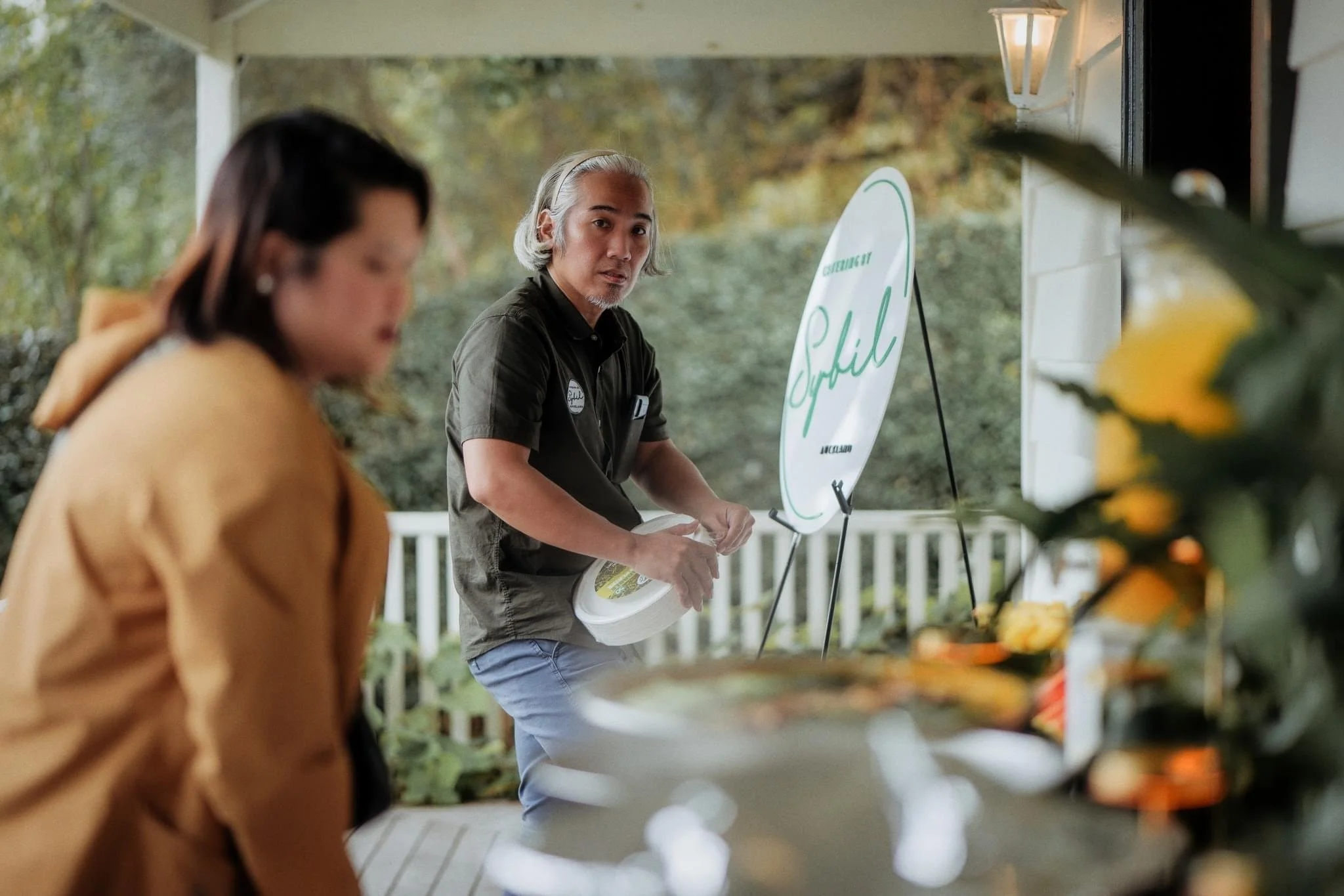 A man with gray hair and a beard, wearing a black shirt, looks at a woman who is out of focus in the foreground. The man is holding a white container, standing outdoors near a table with flowers and a sign that reads 'Social' in green cursive. The ba