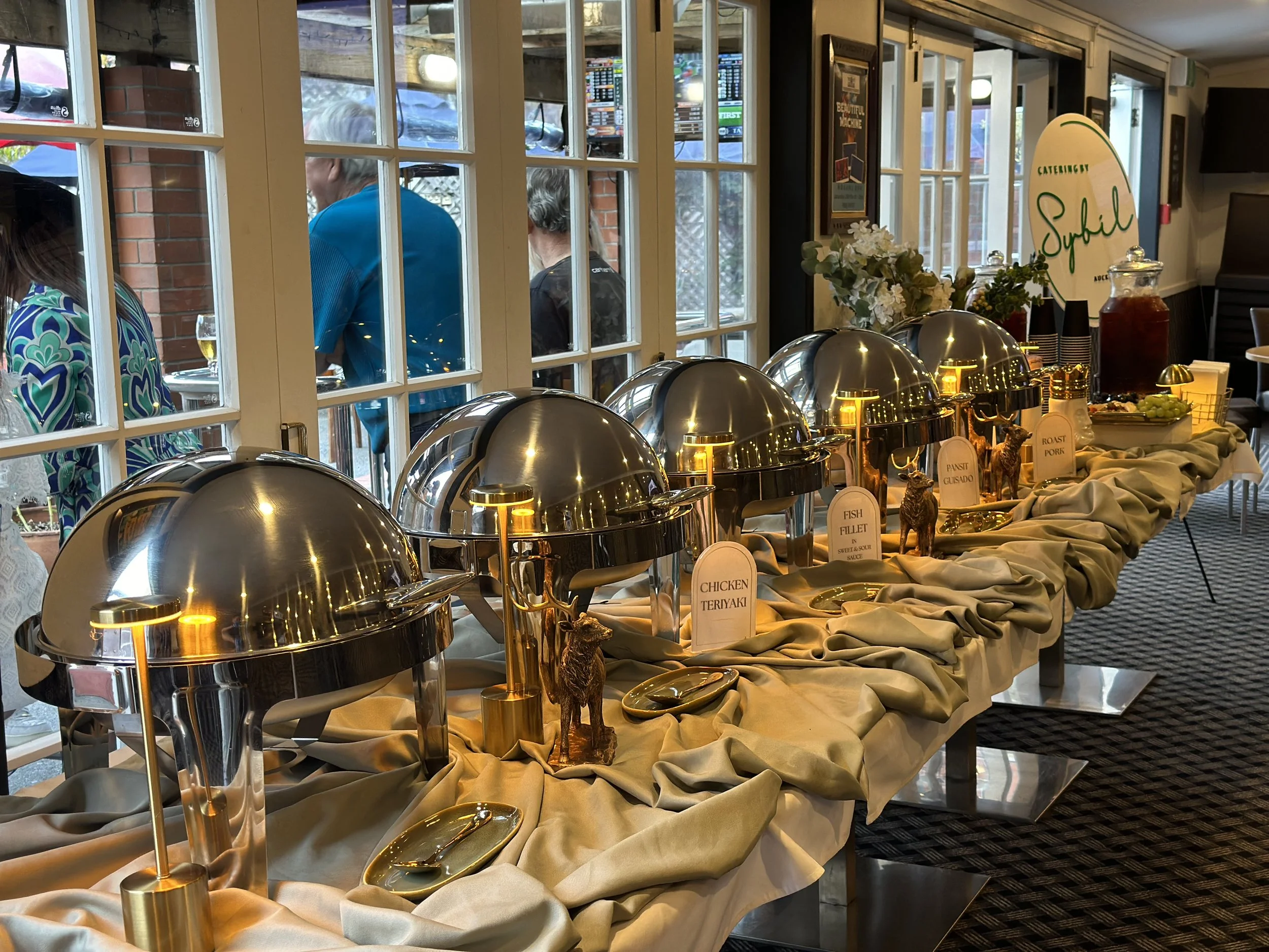 Buffet table with chafing dishes labeled chicken teriyaki, fish fillet, pastrami, roast pork, and other dishes, set up with decorative flowers and a sign reading 'Catering by Sybil'.