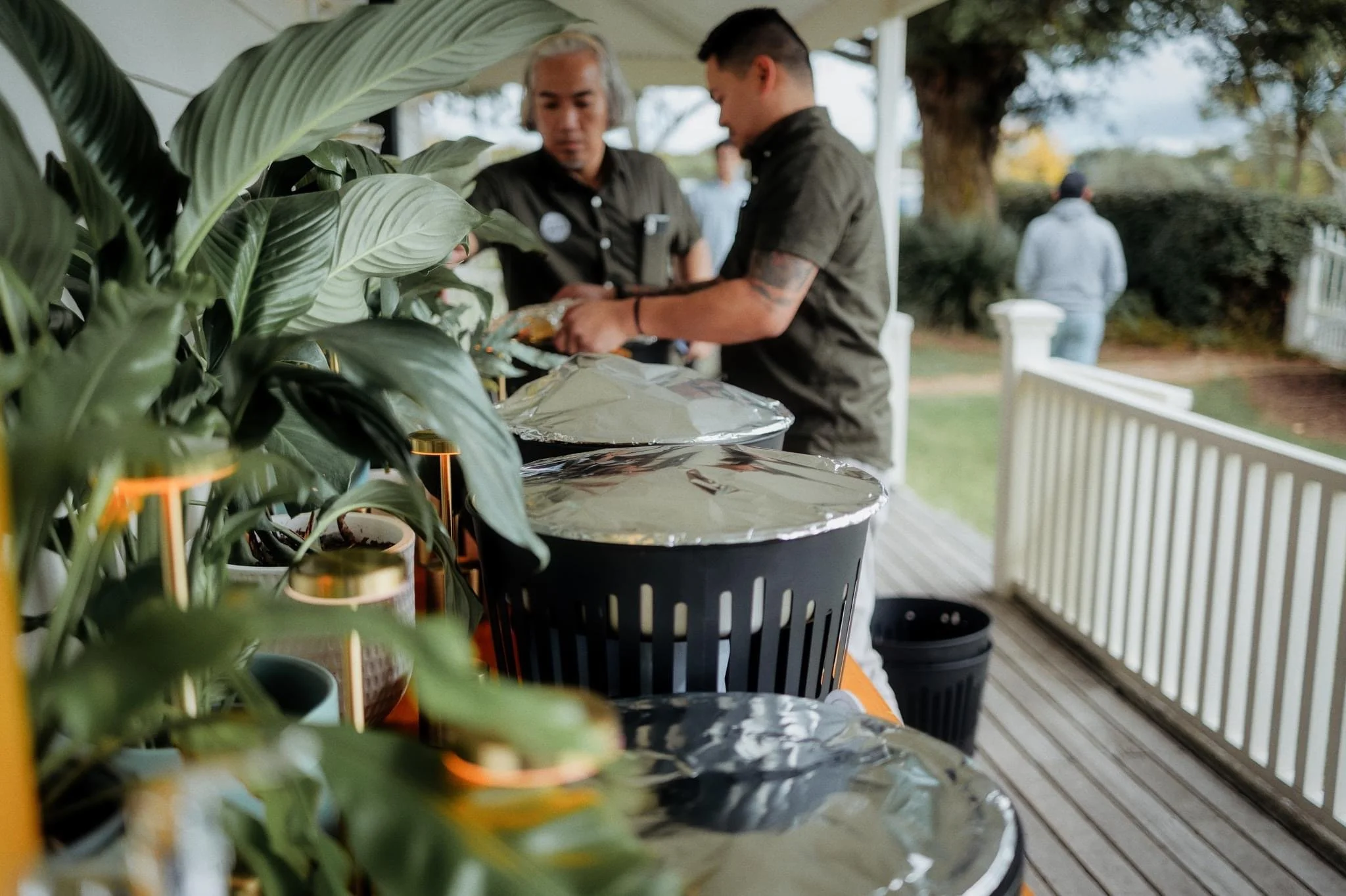 People at an outdoor buffet with covered dishes and plants on a porch.