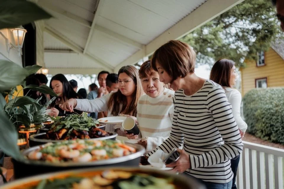People serving and selecting food at an outdoor buffet, with dishes including vegetables and grilled foods on a long table under a canopy.