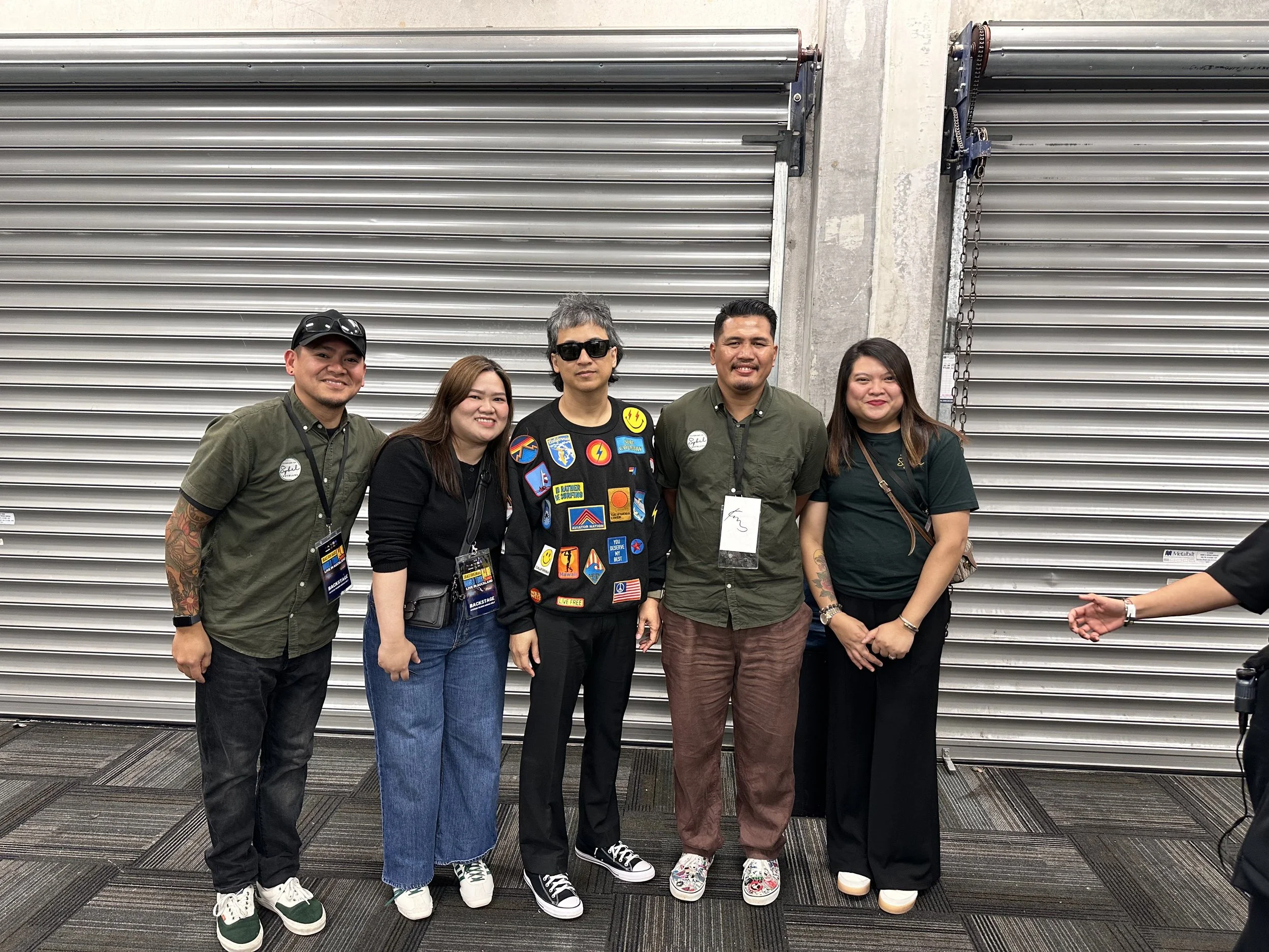 Group of five diverse people standing in front of metallic roller doors at an indoor event, smiling for the camera. Catering by Sybil with E-heads frontman Ely Buendia.