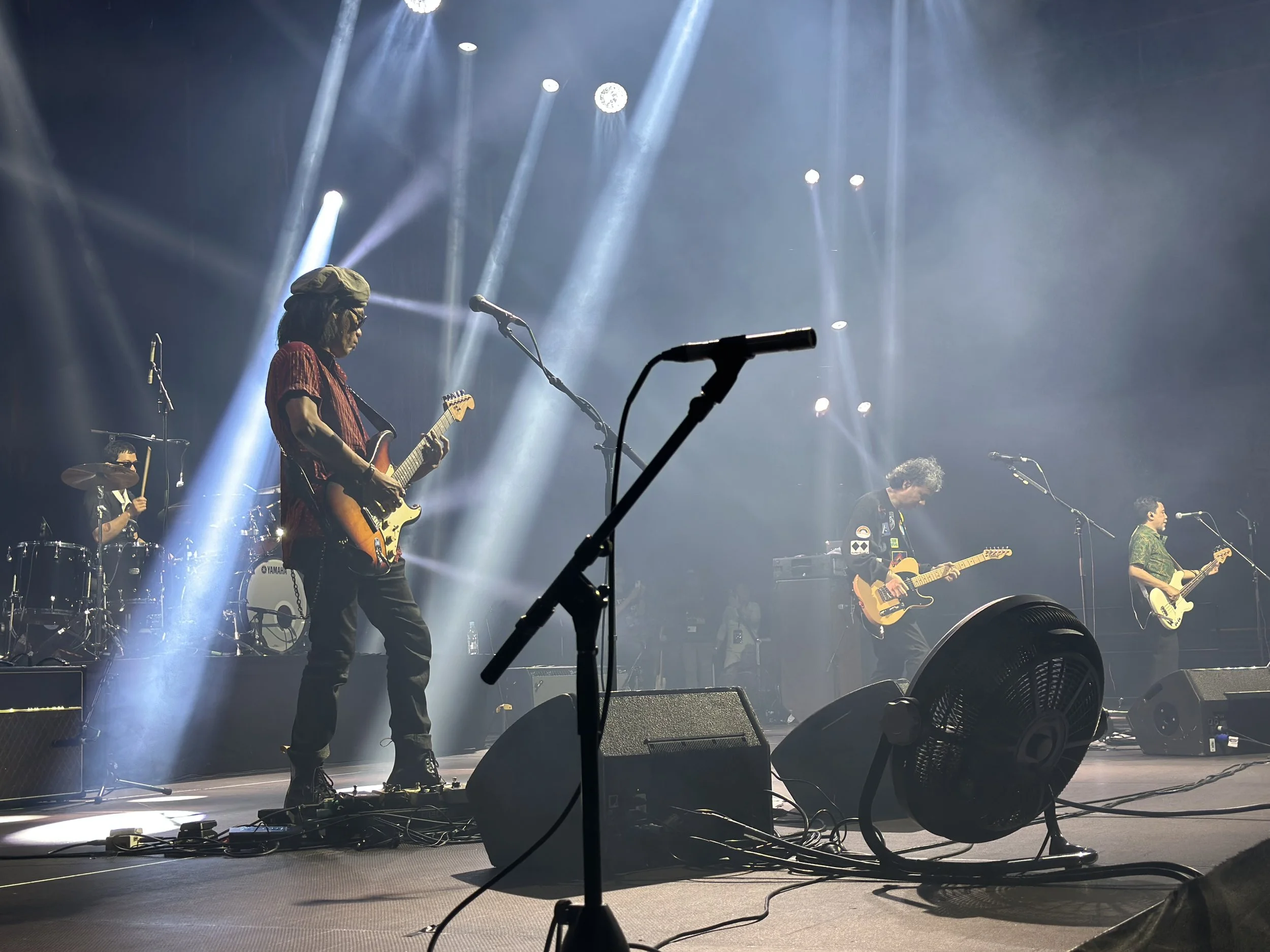 A band performing live on stage with five musicians playing guitars and drums, stage lighting, and sound equipment visible.
