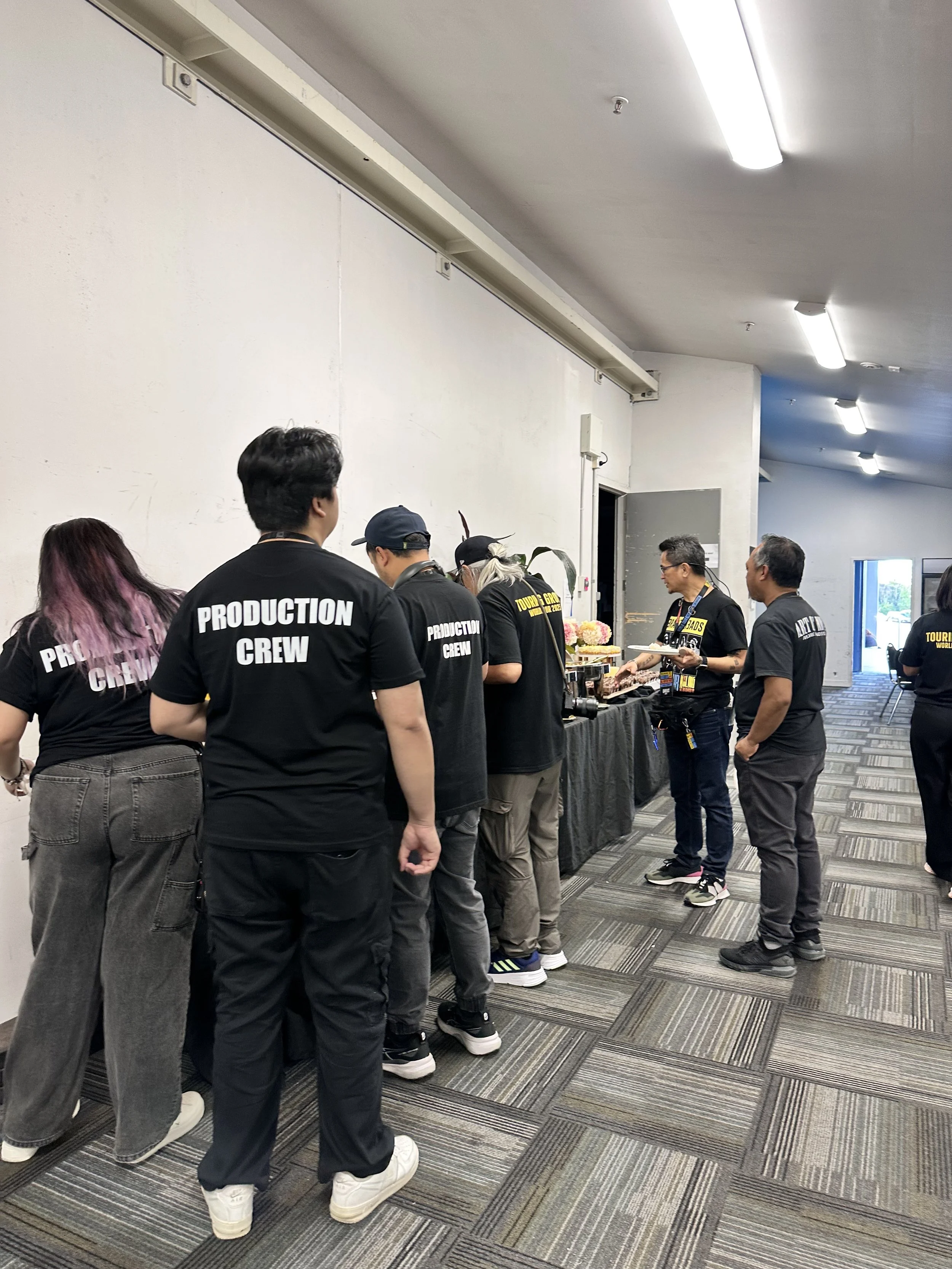 Several people standing in a line at a buffet table in a hallway, wearing black shirts with 'PRODUCTION CREW' printed on the back, serving or waiting for food. This is for Eraserheads concert held in Trust Arena.
