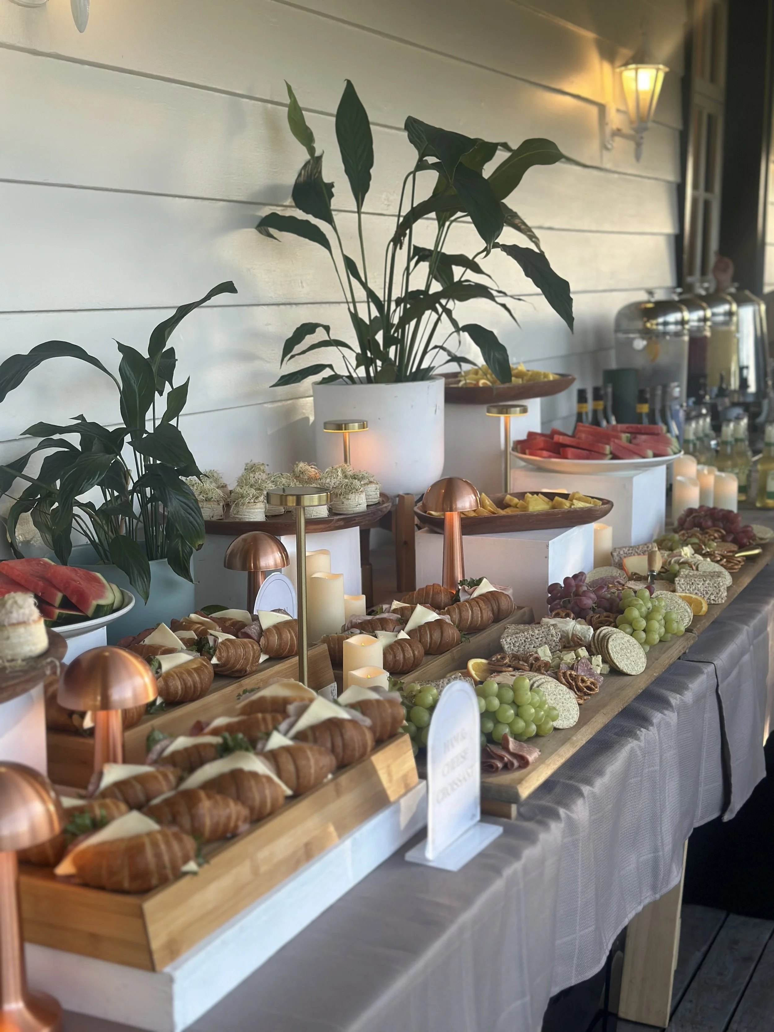Buffet table with assorted fruits, cheeses, pastries, and snacks, decorated with plants and candles, set against a white wall with a lantern light.