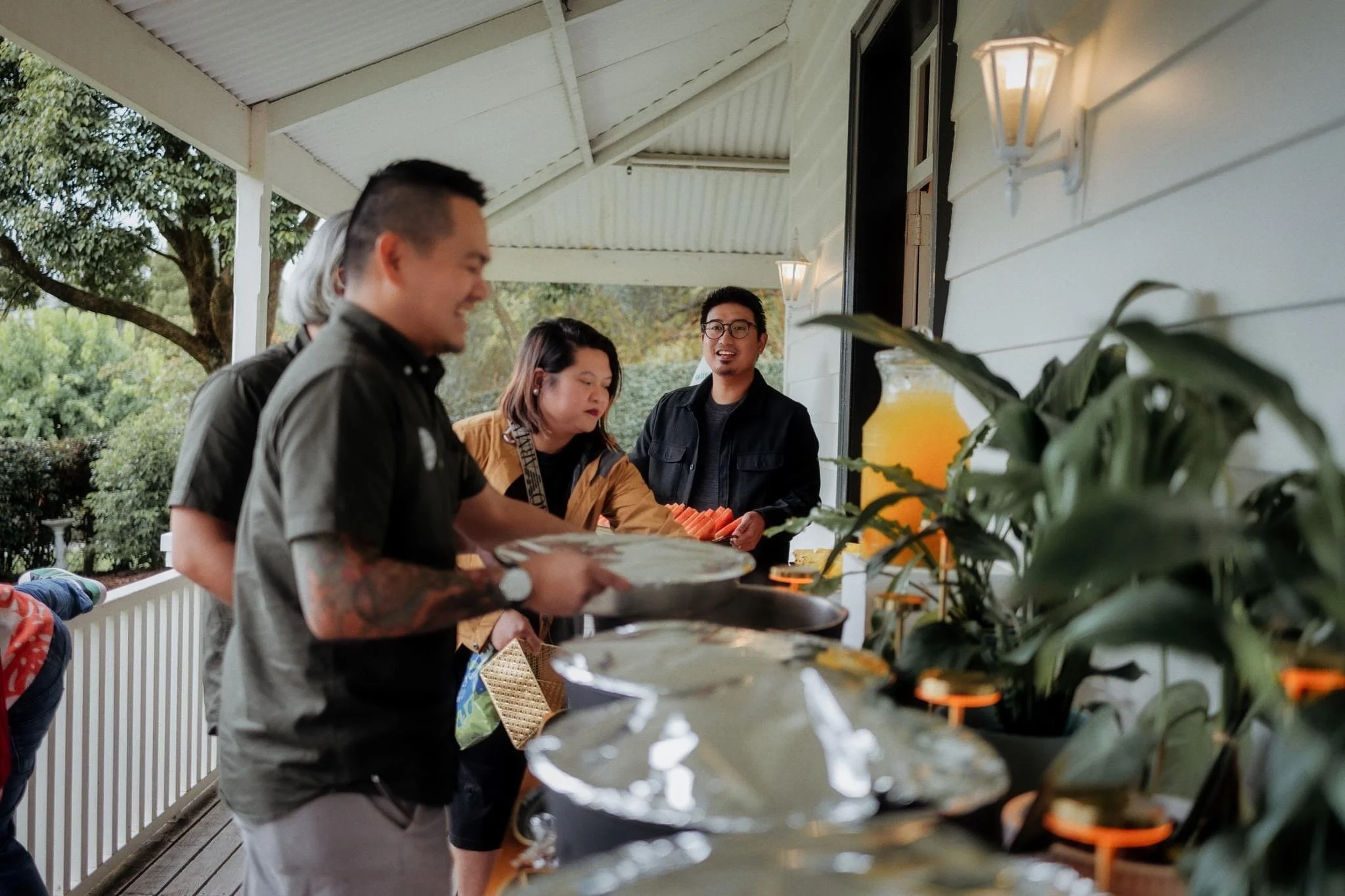 People serving themselves at a buffet on a porch with green trees outside, some holding plates and a woman arranging food on her plate.