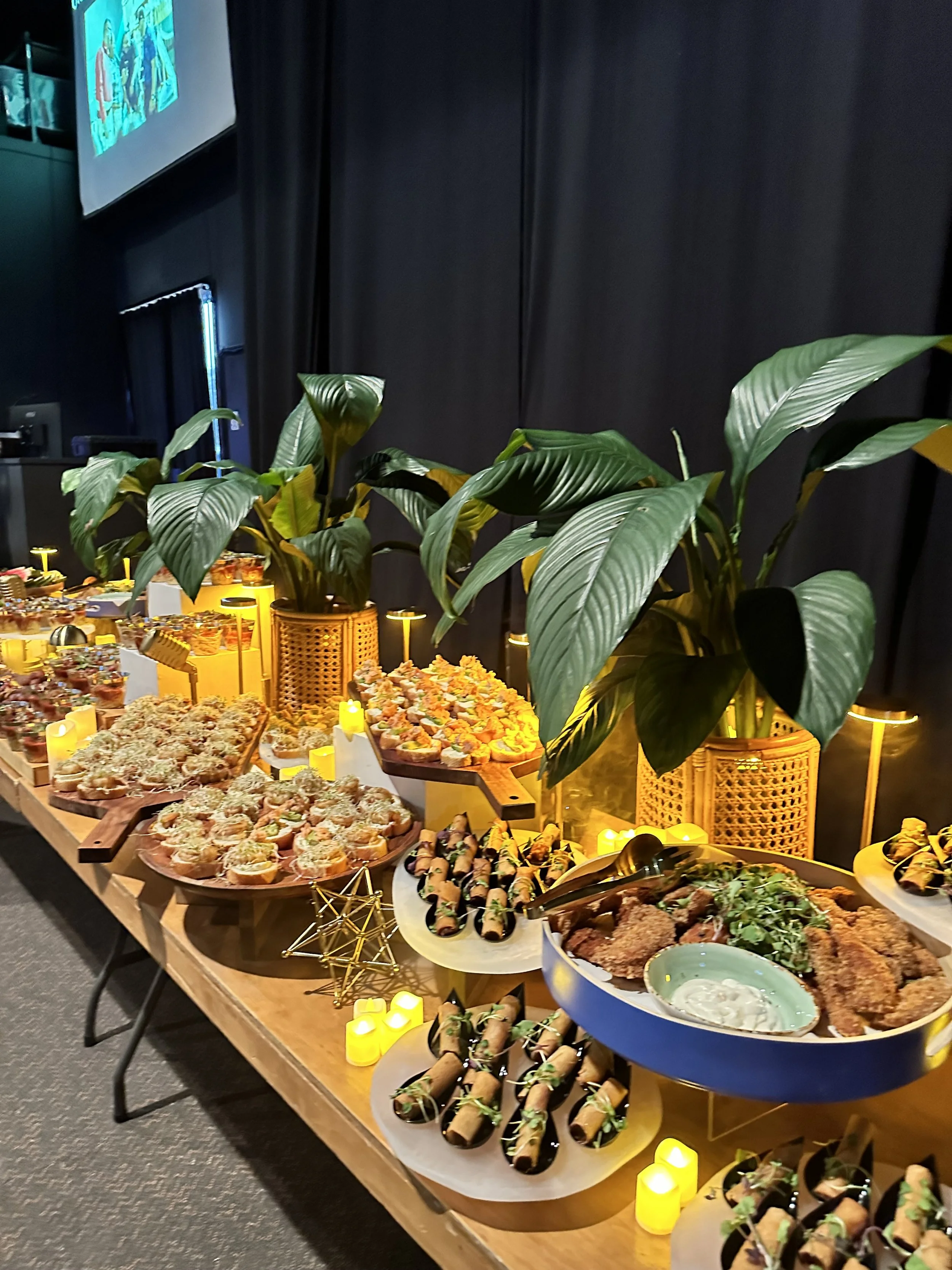A buffet table with various appetizers and finger foods, decorated with large green plants and small yellow candles.