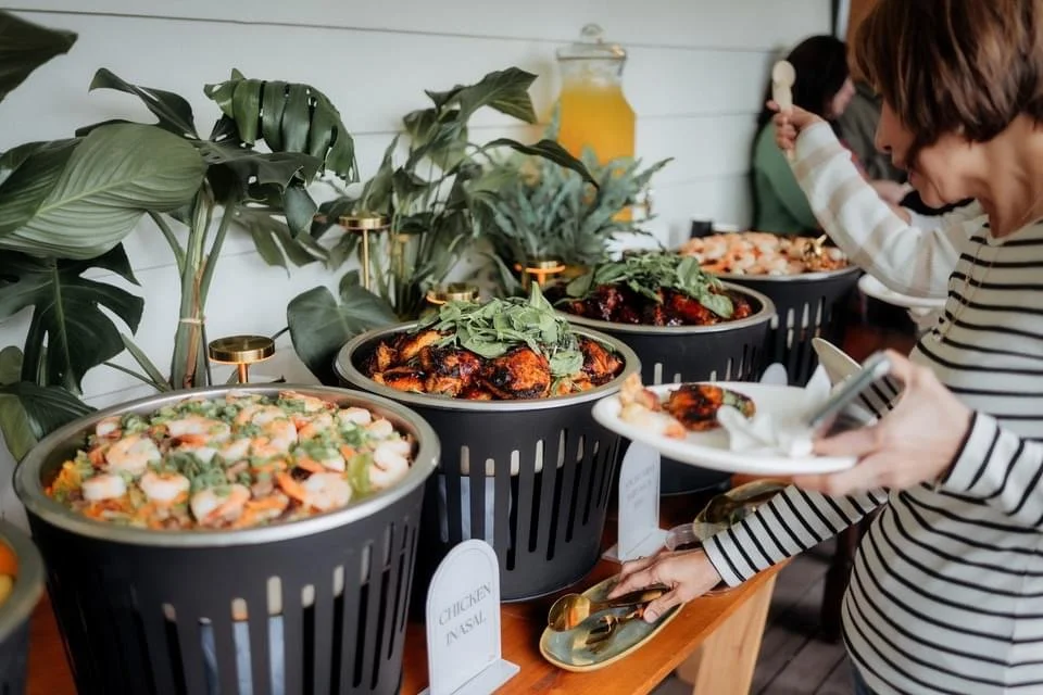 People serving themselves from a buffet of various dishes, including chicken, vegetables, and salads, with greenery and beverages in the background.