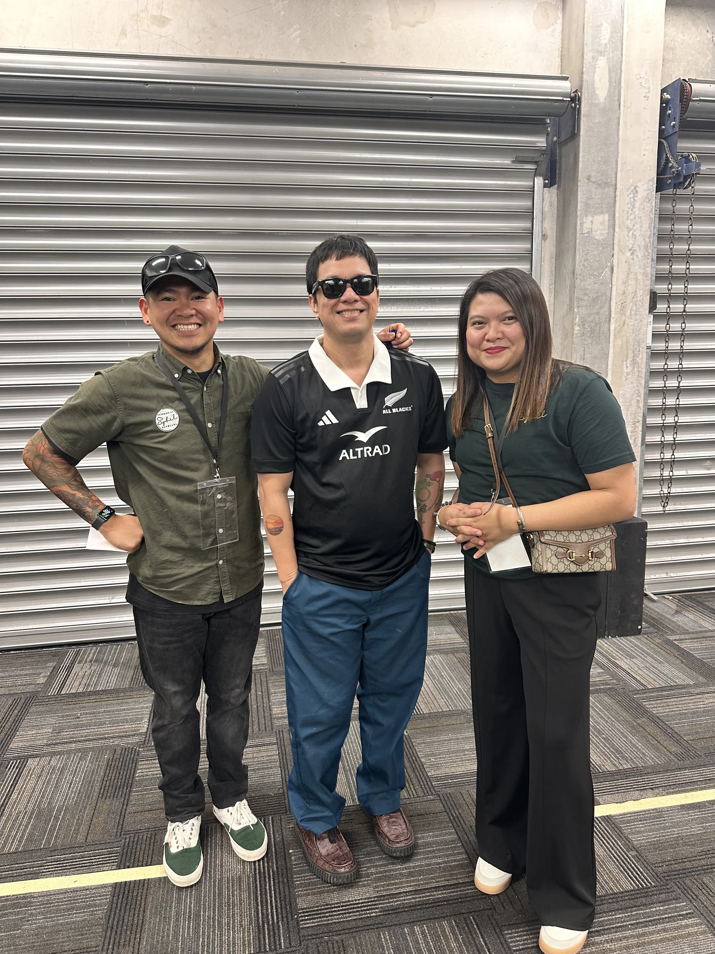 Three people standing together indoors, smiling at the camera. The person in the middle is wearing a black sports jersey with logos, while the other two are in casual attire. Background features a metal roll-up door. CBS with Raymond Marasigan.