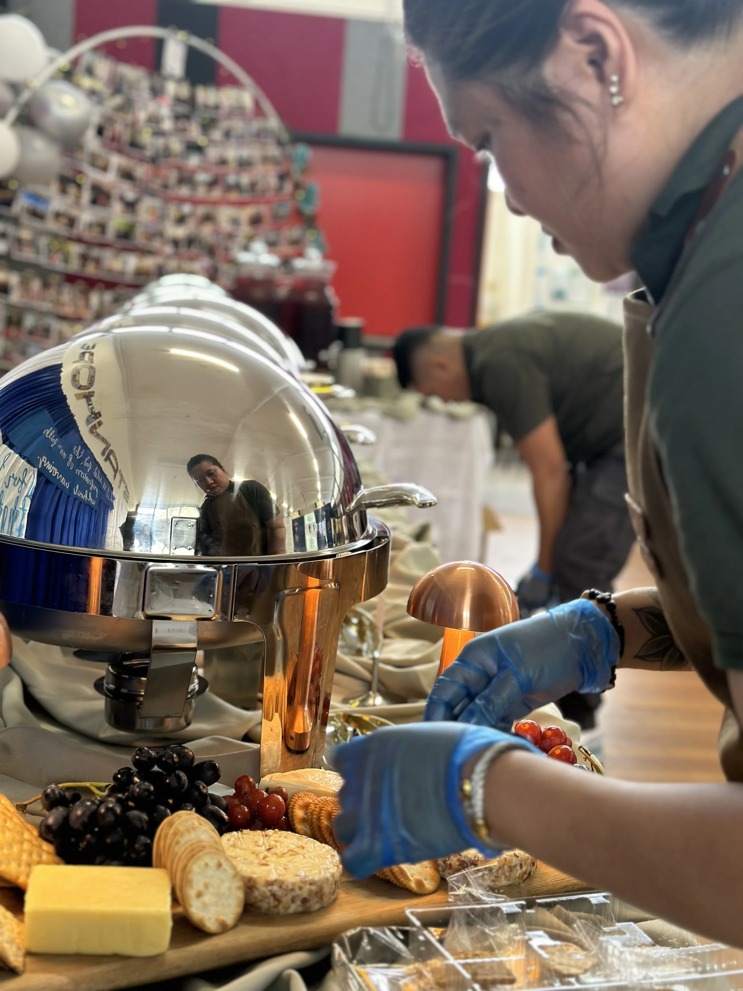 A person wearing blue gloves arranging food on a table with cheese, grapes, crackers, and a chafing dish in an indoor setting, with other people in the background.