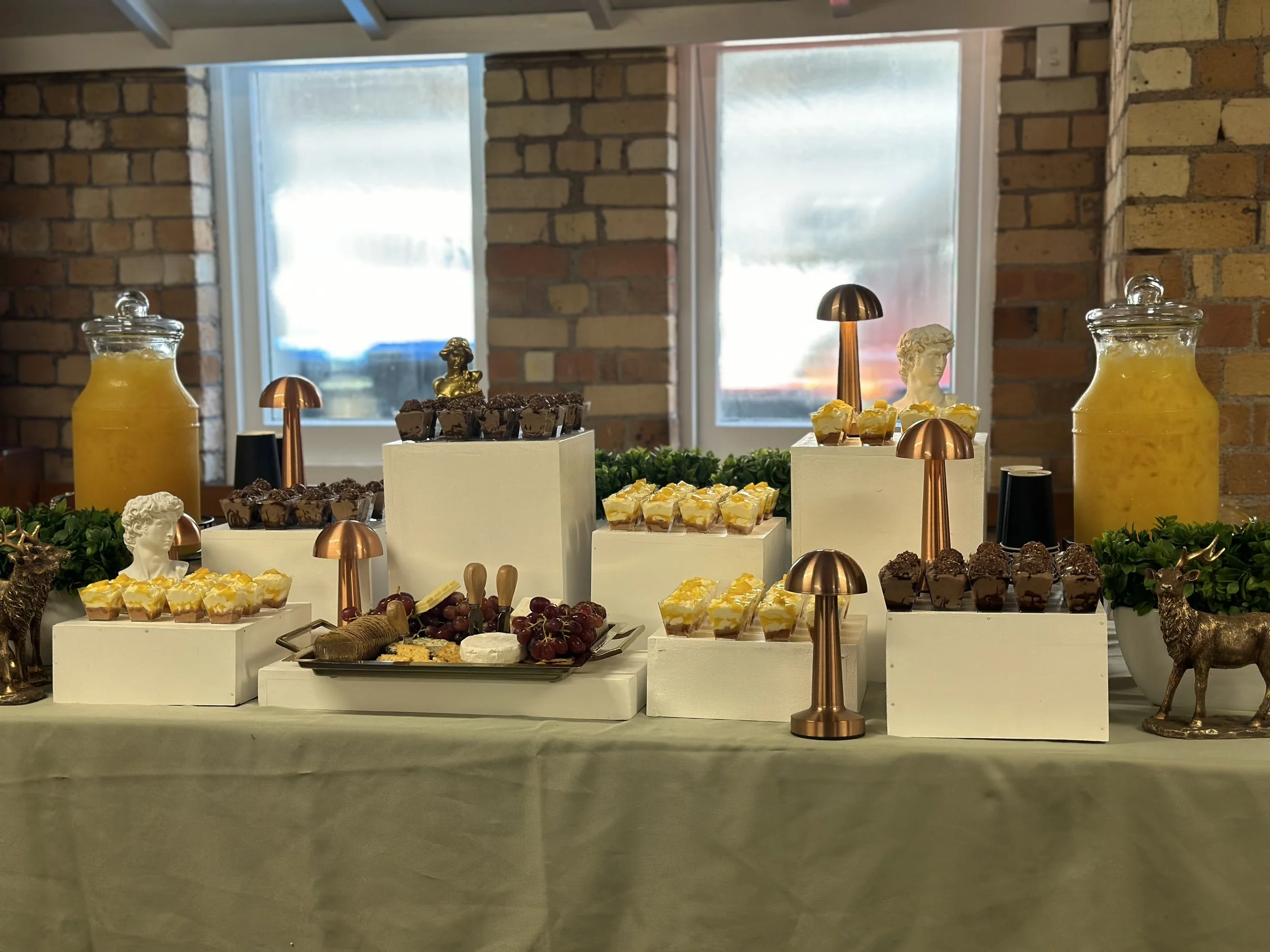 Dessert table with cupcakes, cheese, grapes, and decorative items, set against a brick wall with windows