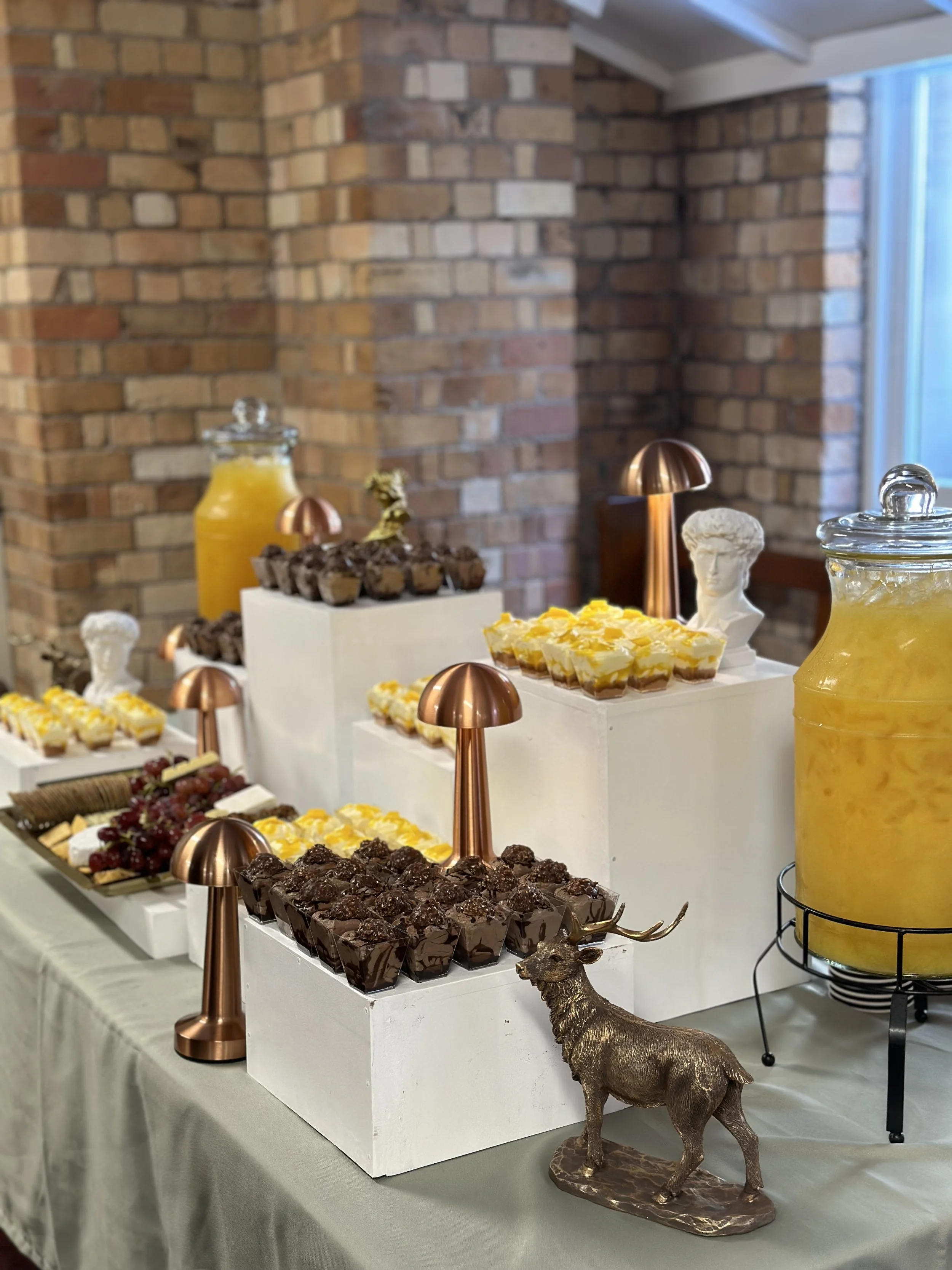 Dessert table with chocolates, cakes, and drinks, decorated with small bronze mushroom ornaments and a bronze reindeer figurine, set against a brick wall.
