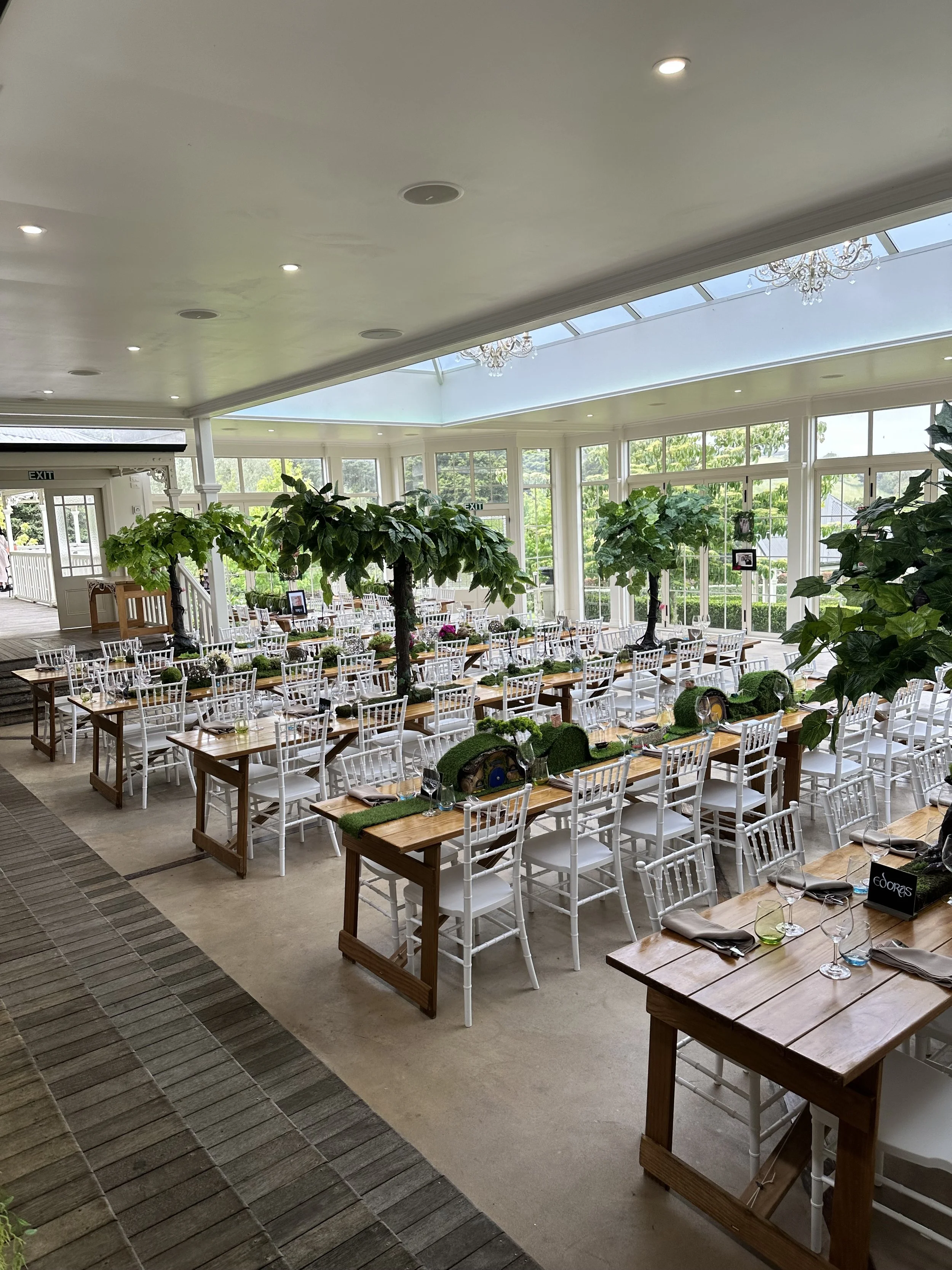 A beautifully decorated indoor dining area with wooden tables, white chairs, and large decorative trees in the center. The space has large windows and a glass ceiling, allowing lots of natural light. The tables are set with glassware and napkins, arr