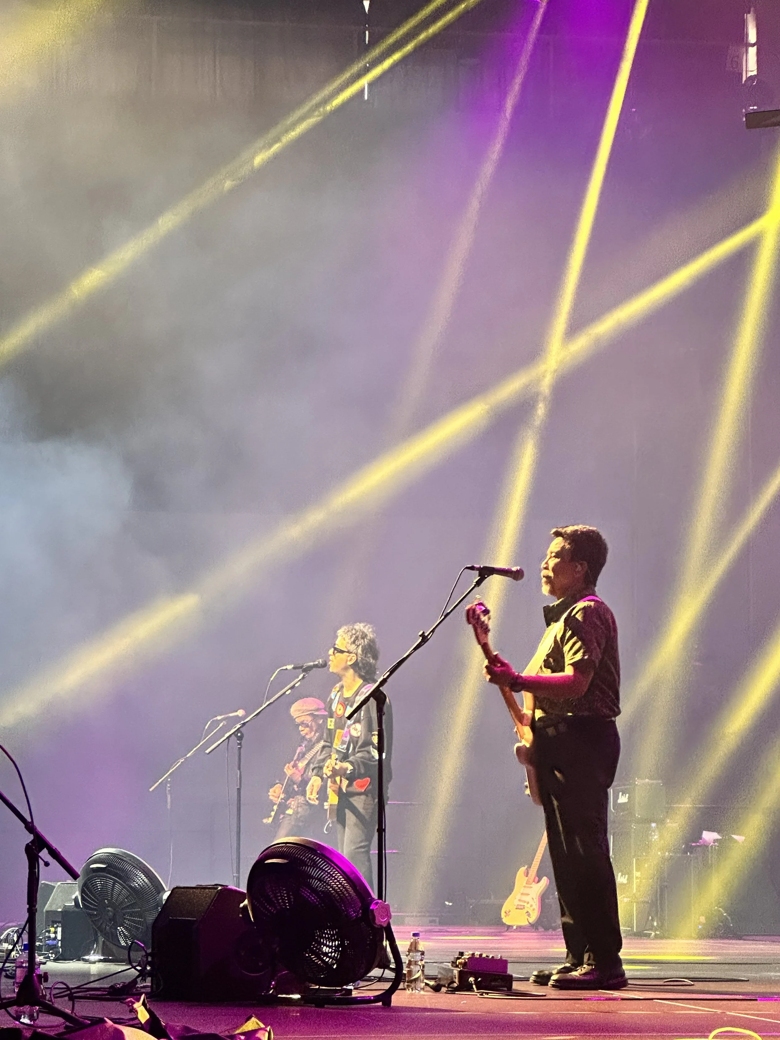 Smoky stage with yellow and purple lighting, band members playing guitars and singing into microphones.