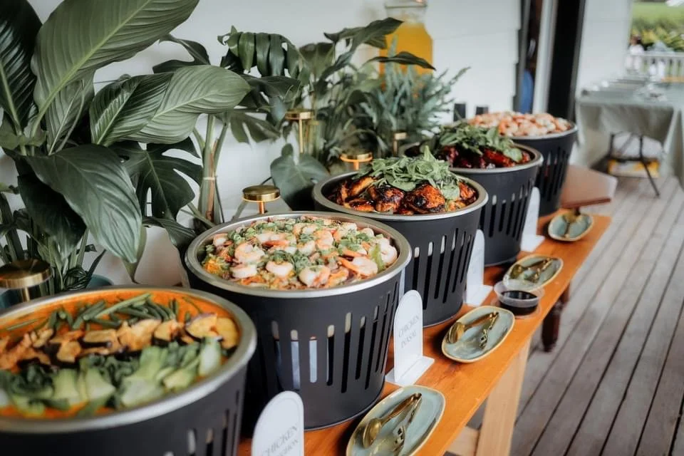 A row of four black ceramic bowls filled with various salads on a wooden table, with green houseplants in the background.