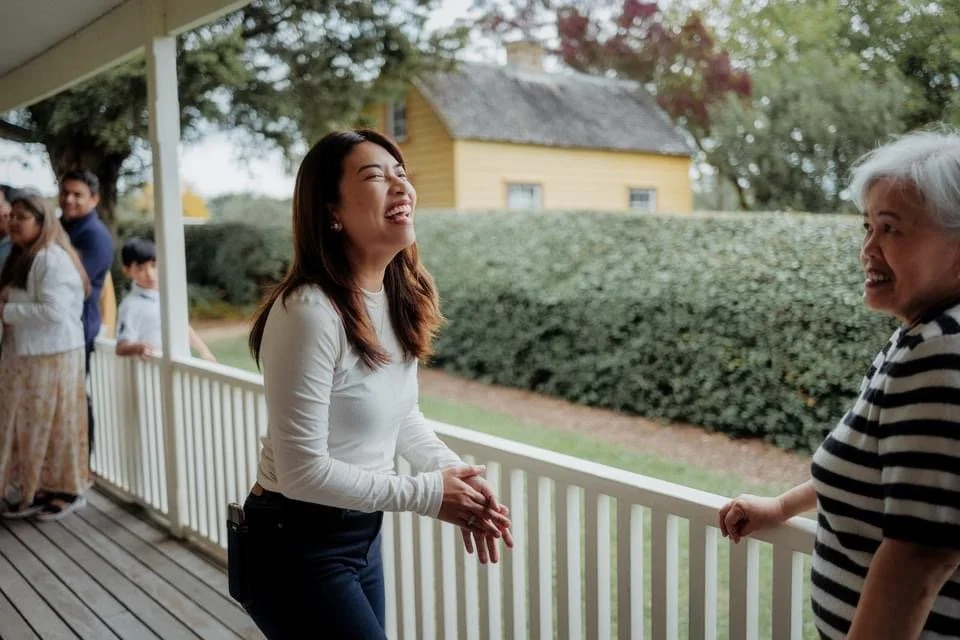 Two women are smiling and talking on a porch, with a group of people in the background, and a yellow house with bushes and trees behind them.