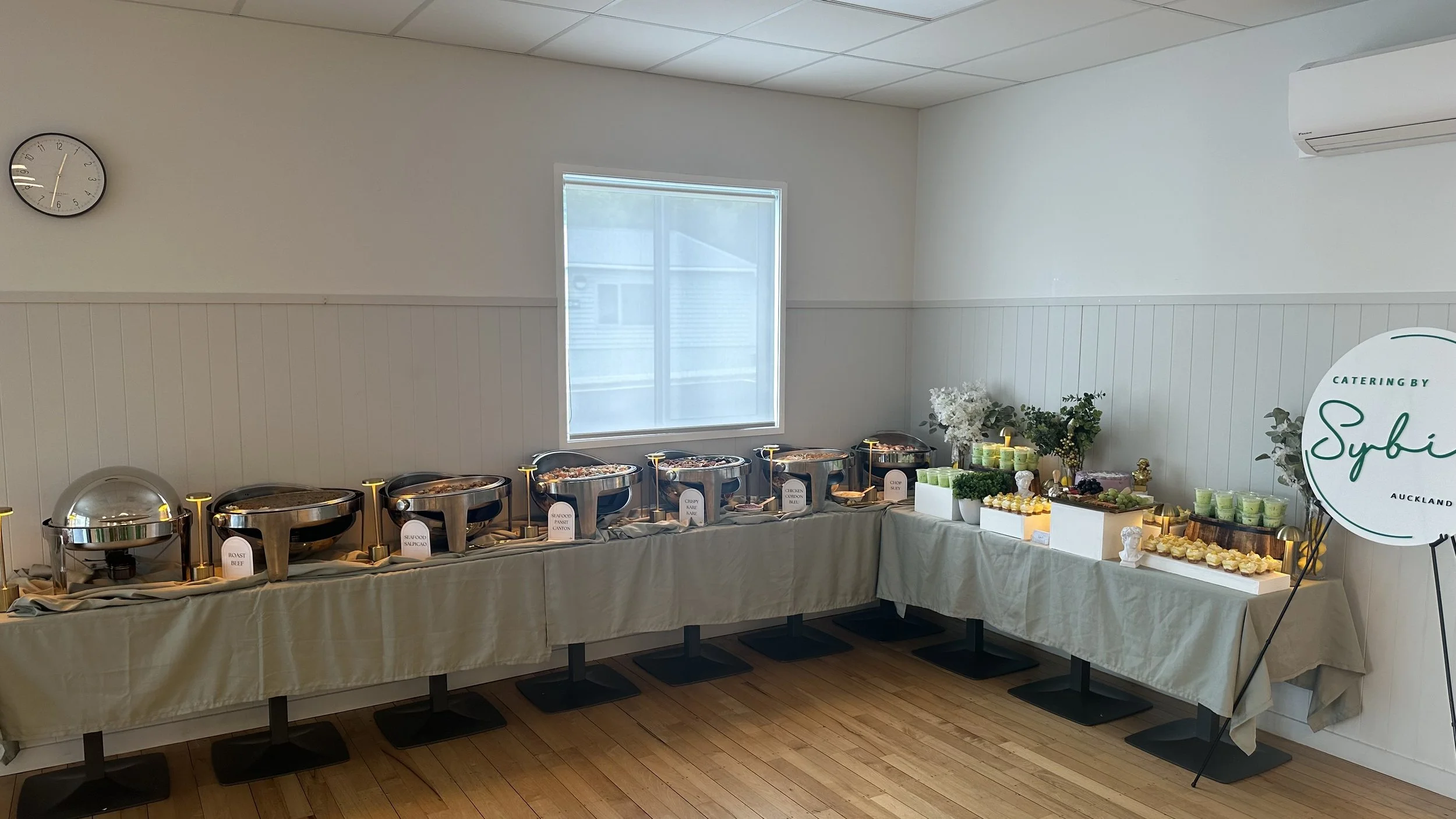 Buffet table with chafing dishes in a bright room, decorated with flowers and refreshments, featuring a catering sign that reads 'Sybi Catering Auckland'.