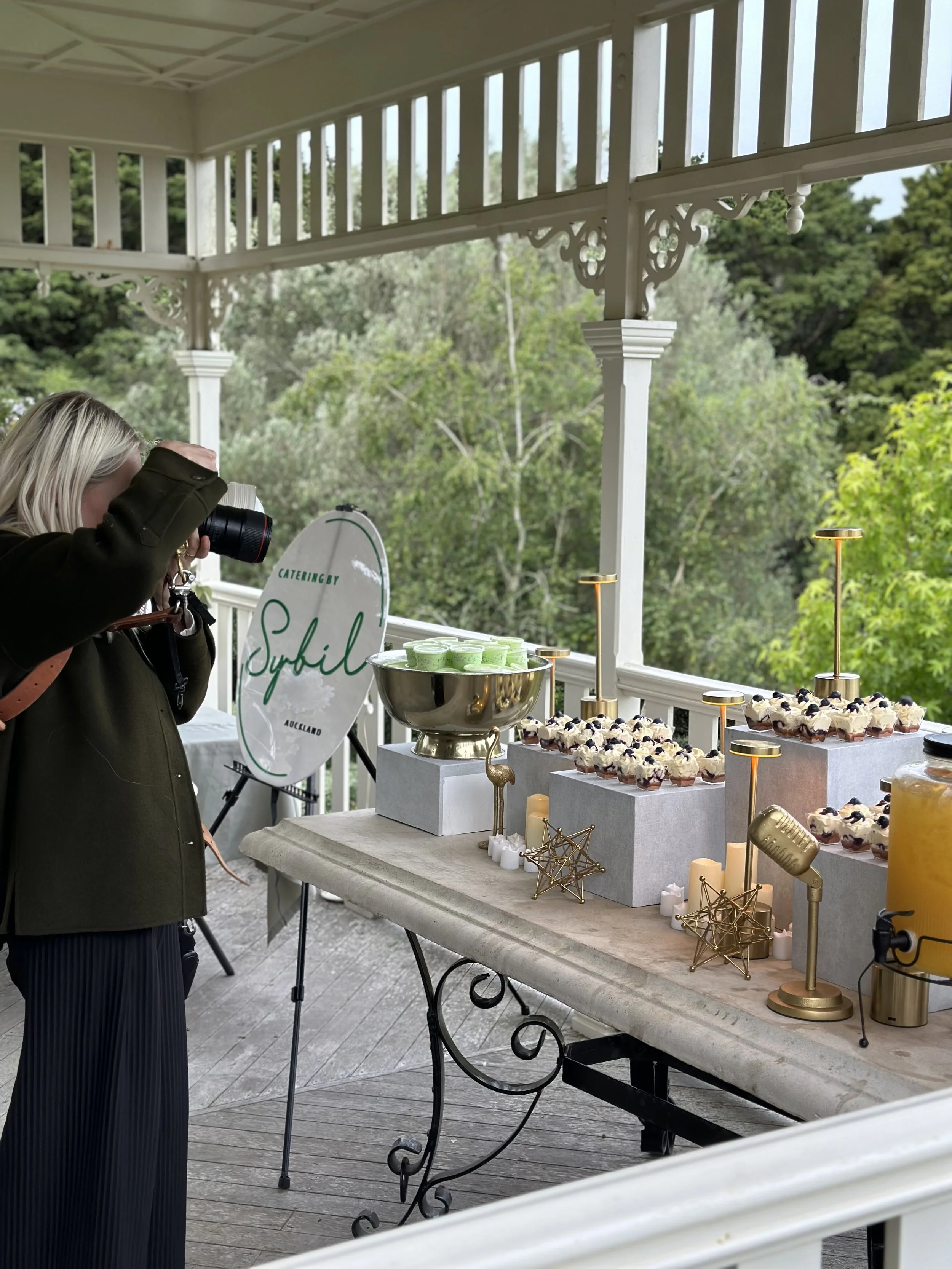Person taking a photo of a dessert table at an outdoor event with cupcakes, candles, and a sign that says "Catering by Sybil" on a porch surrounded by trees.