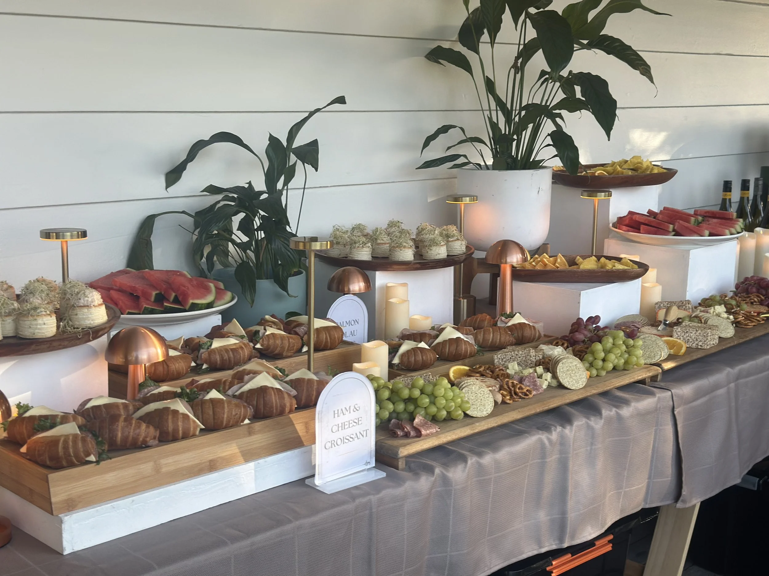 A buffet table with an assortment of finger foods including croissants, cheese, grapes, watermelon, sliced pineapples, and assorted meats. The table is decorated with small lamps, candles, and green plants in white pots.