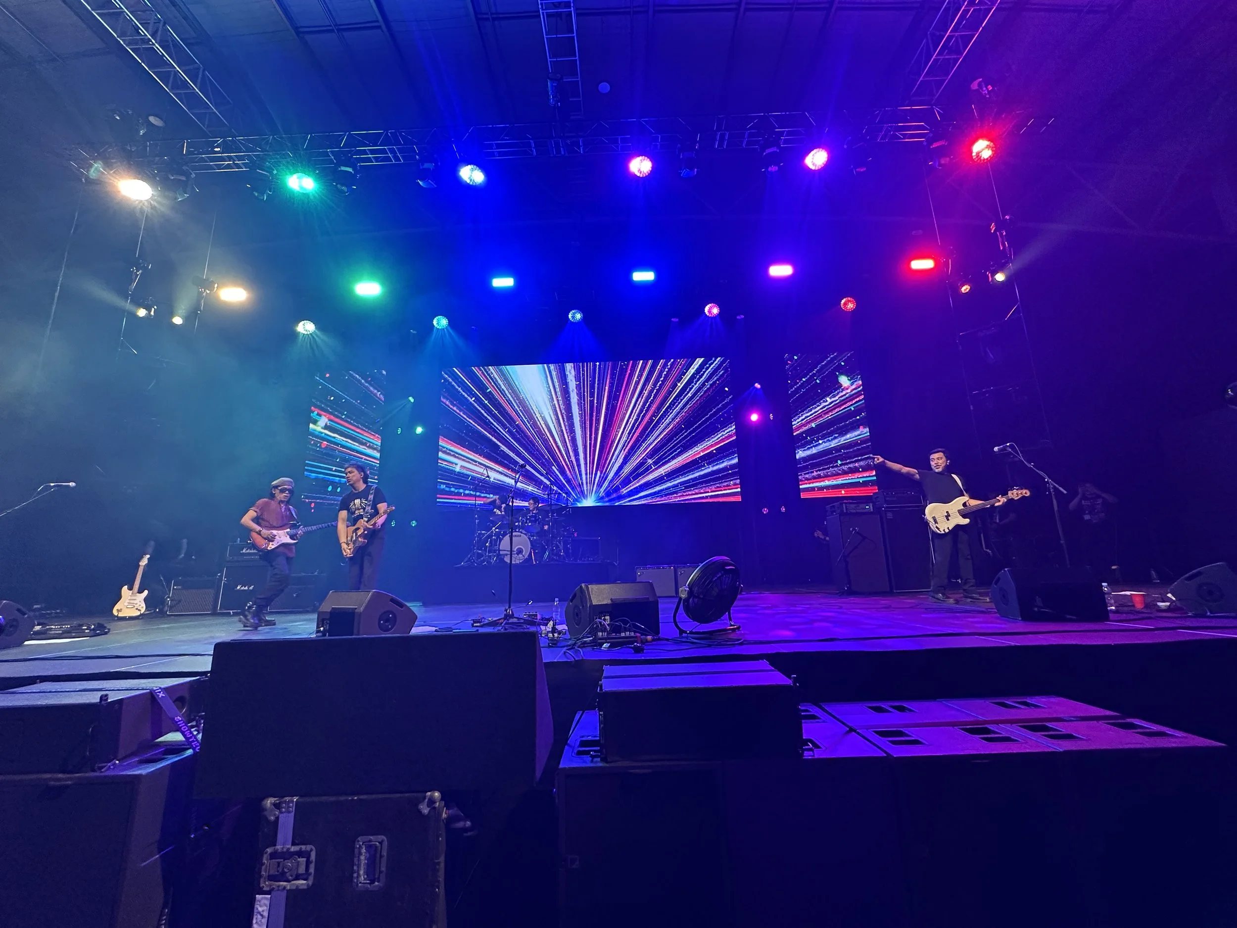 A live band performs on stage with colorful stage lights and a digital backdrop displaying a starburst pattern.