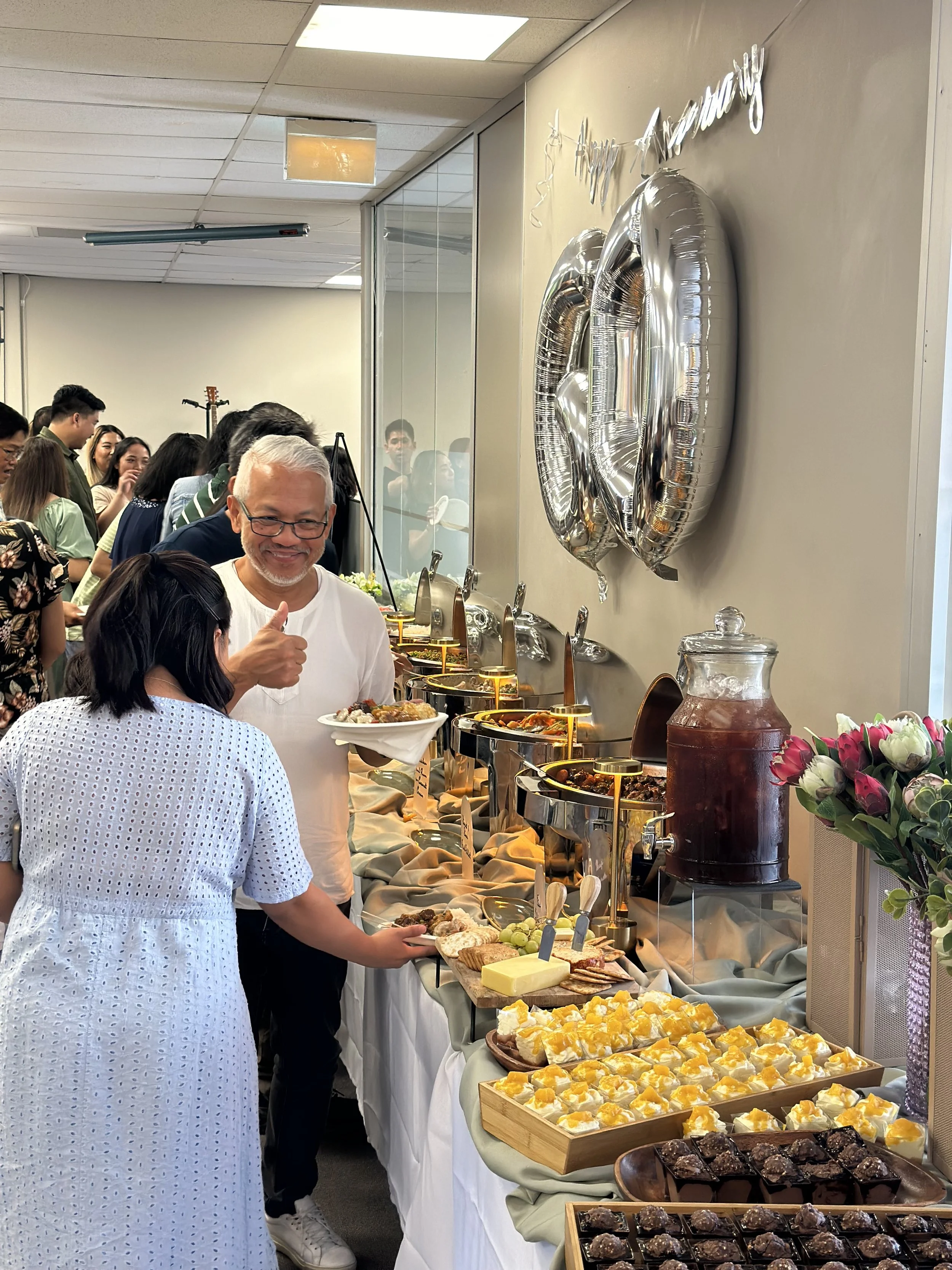 A buffet table at a celebration with various desserts and drinks, with people serving themselves and a man giving a thumbs-up.