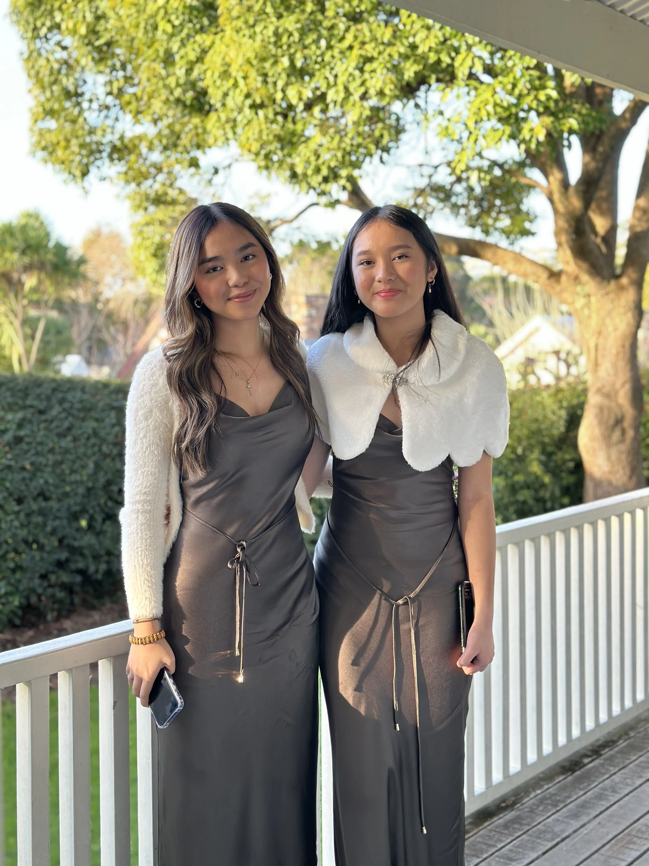 Two young women standing outdoors on a porch, dressed in elegant black dresses with white shawls, smiling at the camera, with trees and a clear sky in the background.