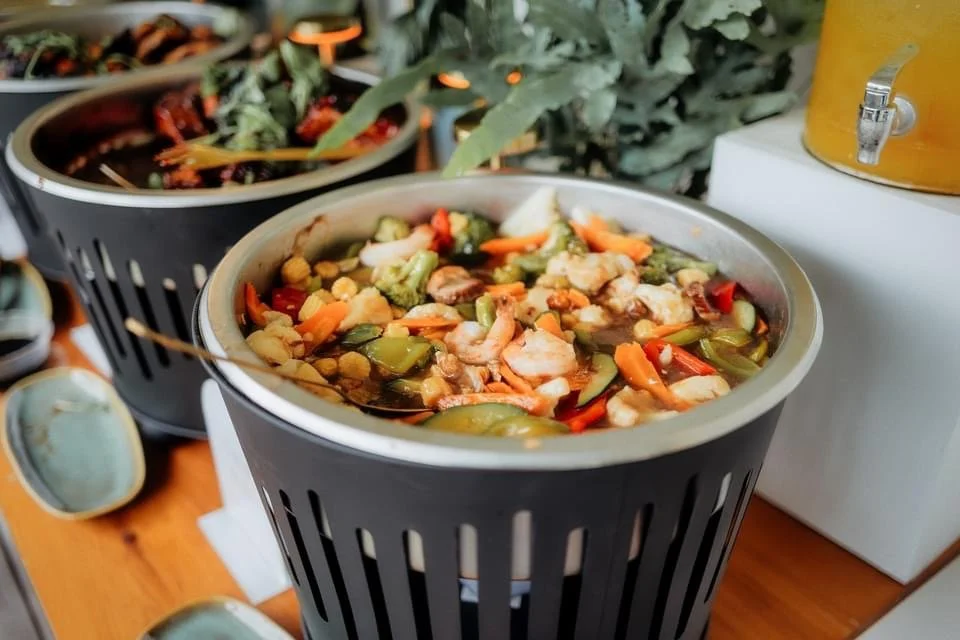 A large pot of vegetable and chicken soup on a buffet table, with other dishes and greenery in the background.