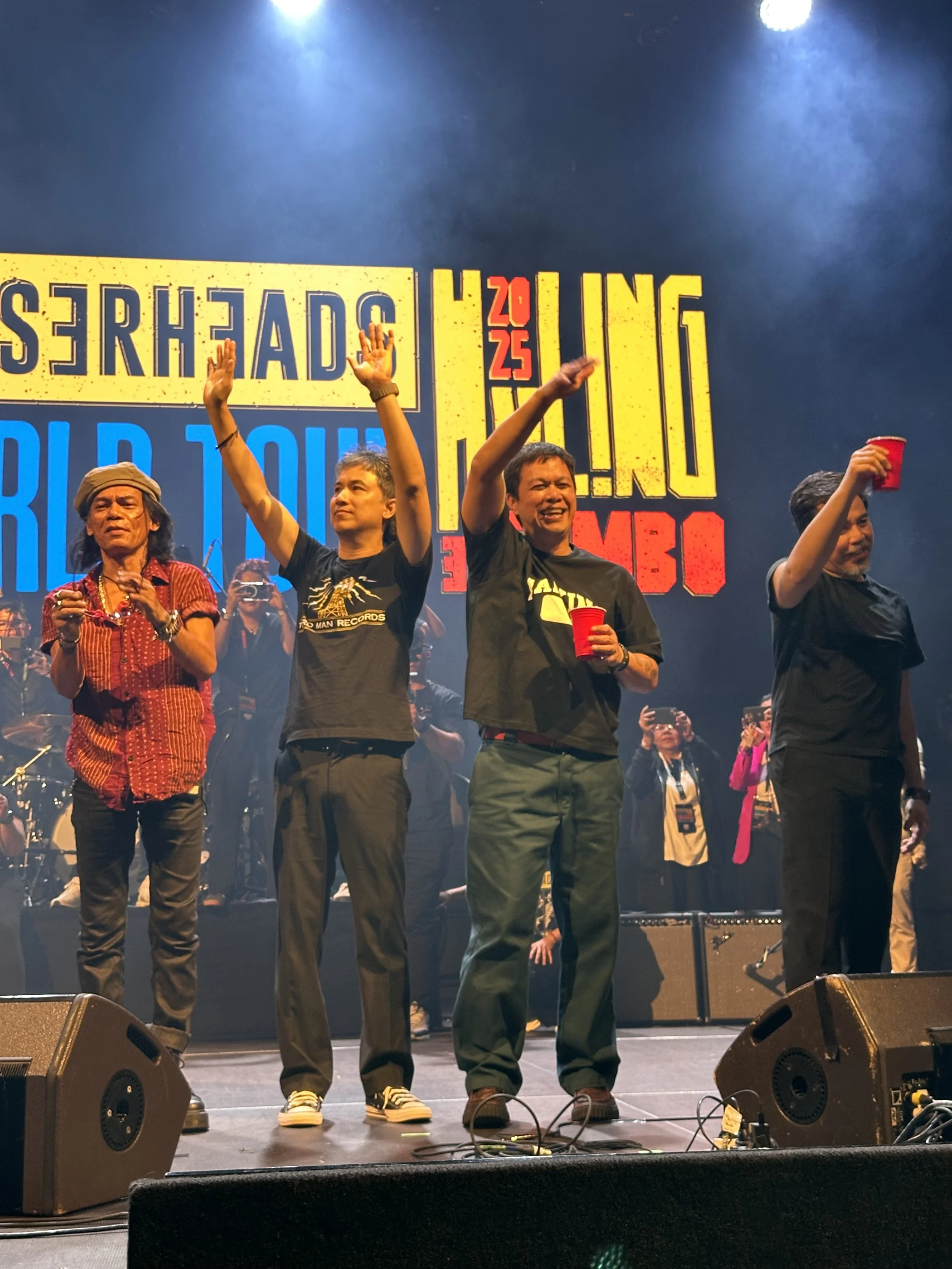 Eraserheads on stage at their concert, holding red cups, with three of them waving or raising their hands. Behind them is a large screen with colorful text and graphics, and there are other people in the background taking photos or videos.