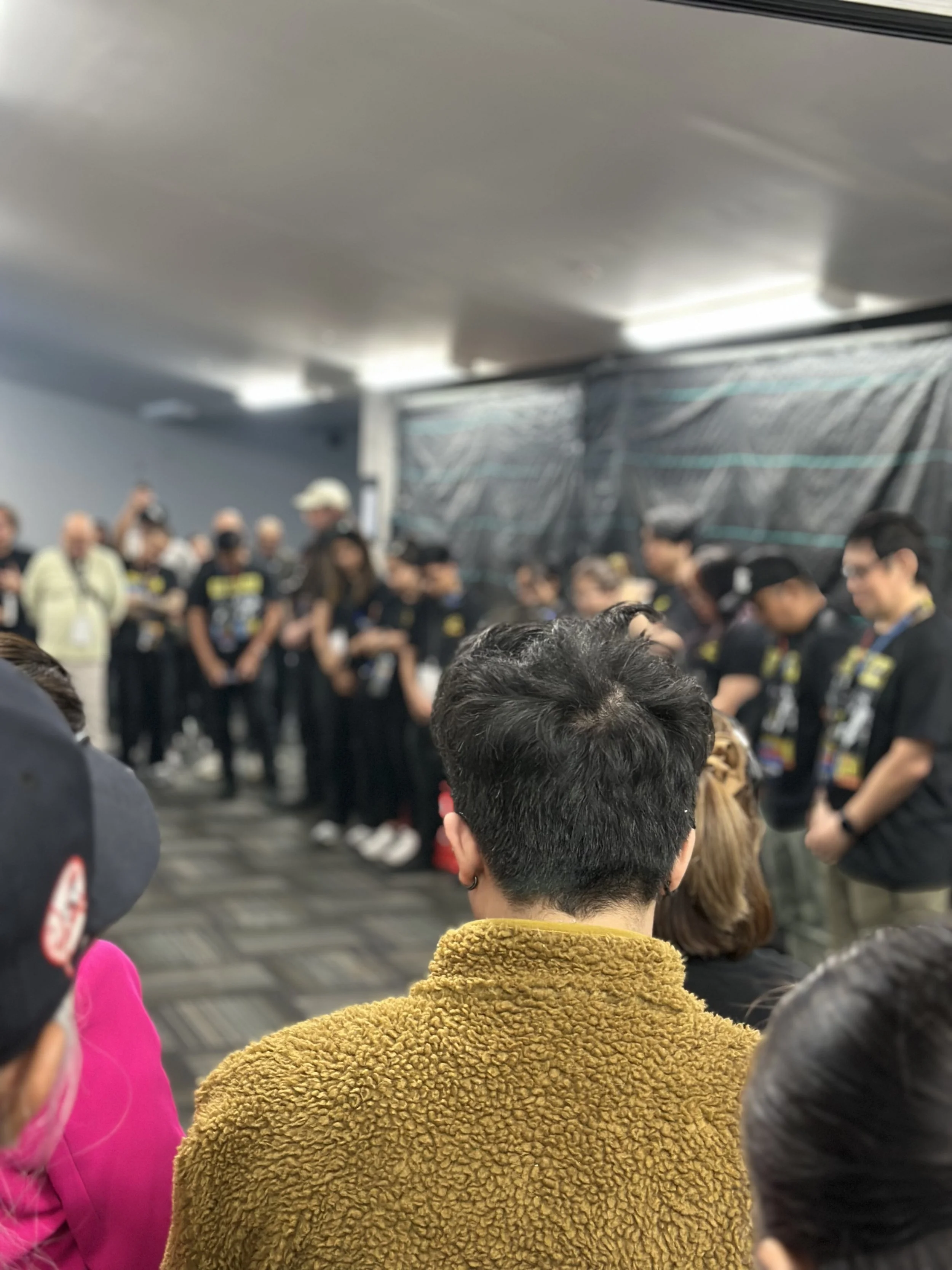 People gathered in a crowded indoor space, some standing in line, with a black backdrop and blurred background, and the photo is taken from behind a person wearing a mustard-colored fleece jacket. Eraserheads pre concert prayers.