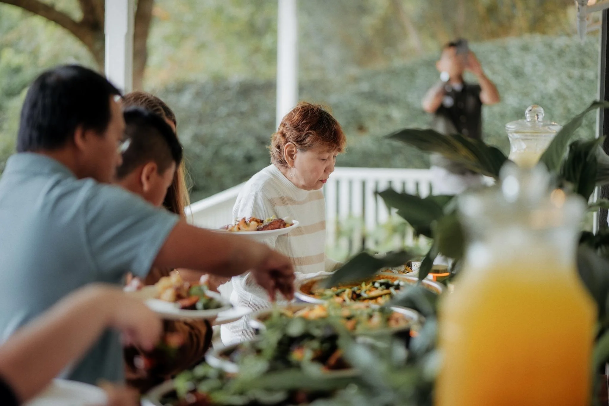 People serving themselves food at an outdoor buffet table with a woman in a white striped sweater in the center, with a man and others on the left, and a blurred person taking photos in the background.