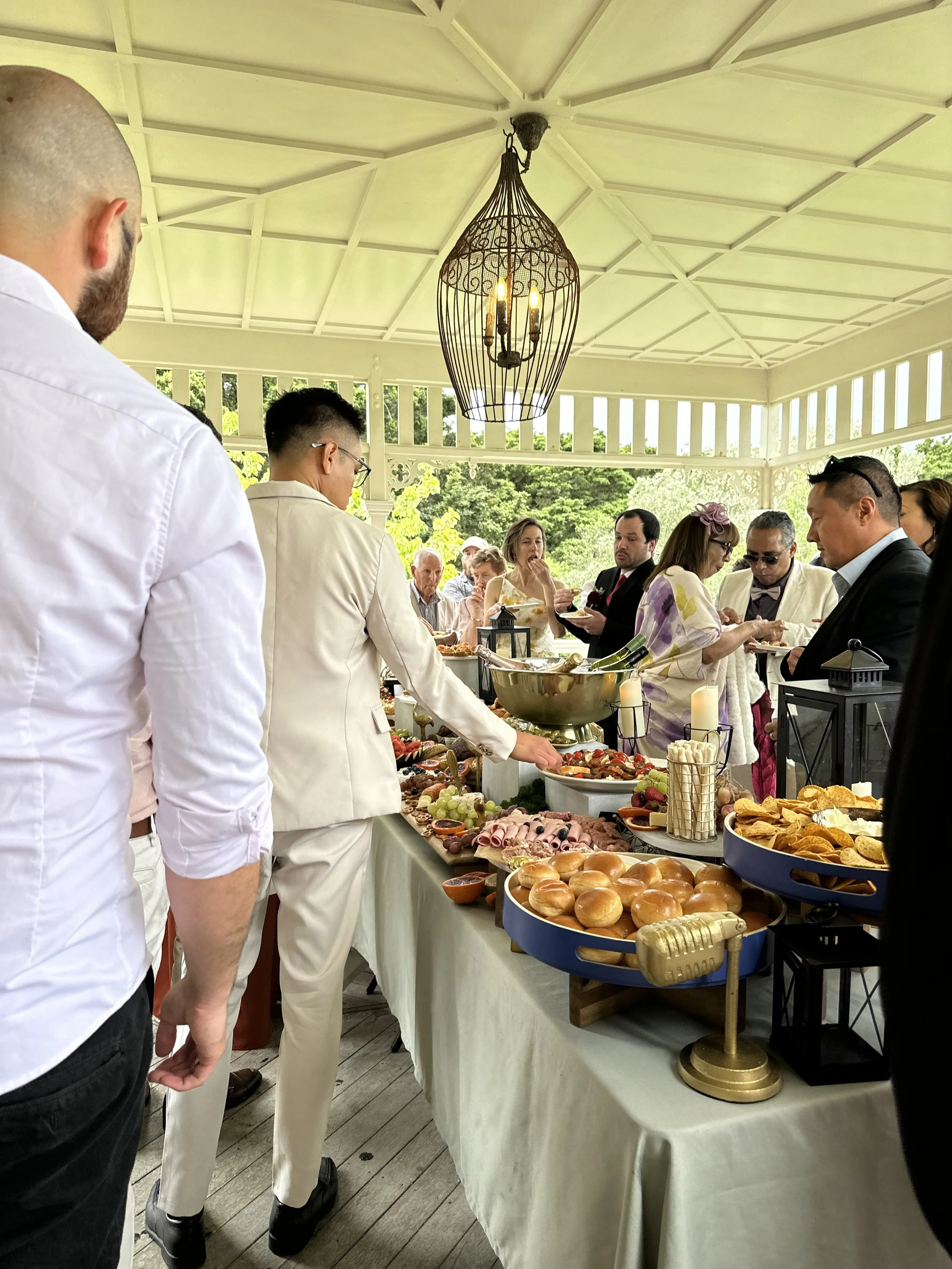 People gathered around a buffet table at an outdoor event, with various foods like bread rolls, meats, cheese, and fruits on the table under a pavilion with a chandelier.