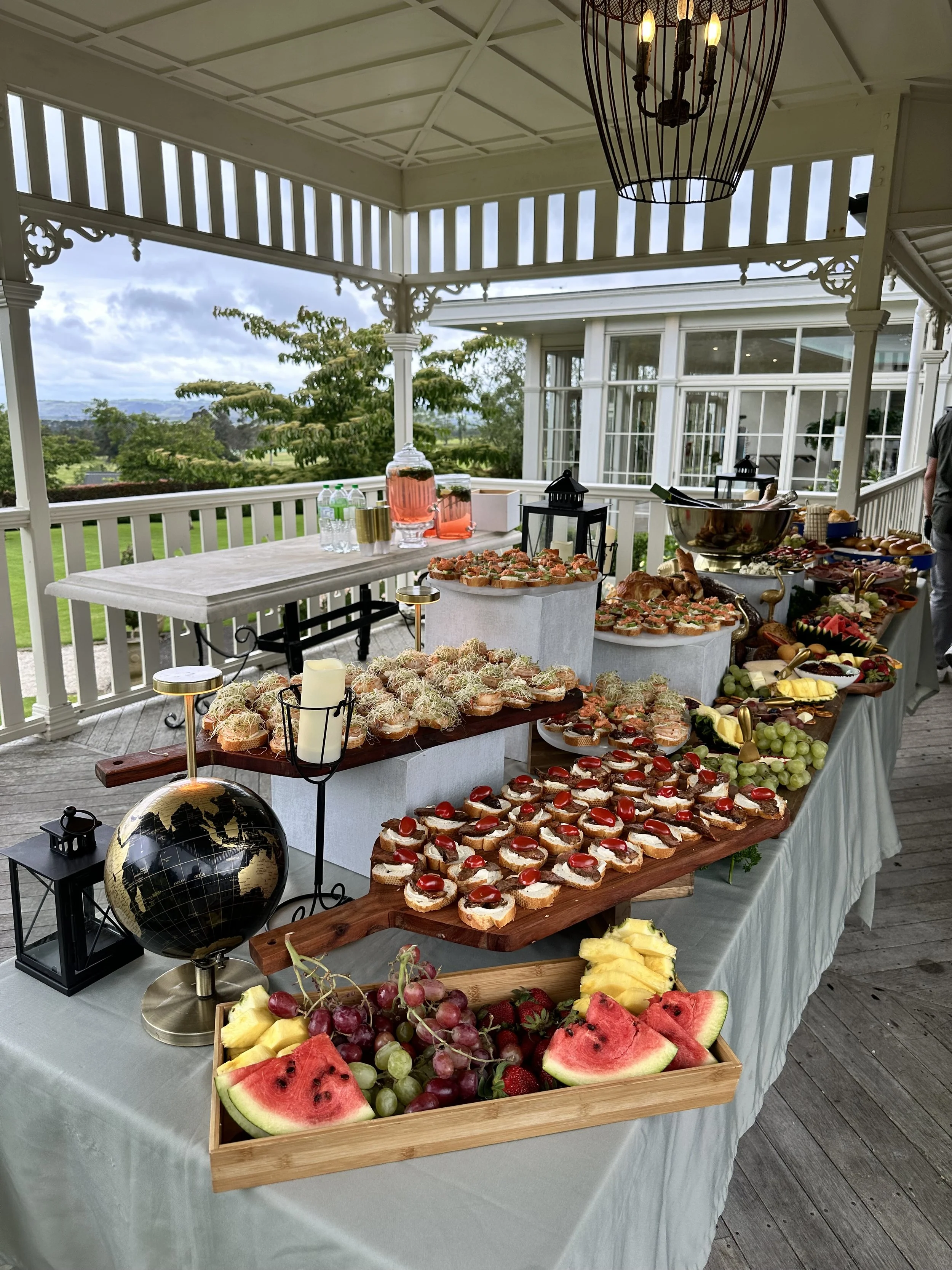 A buffet table with assorted desserts and fruits, including cupcakes, watermelon slices, grapes, and strawberries, set outdoors on a covered porch with a scenic view.