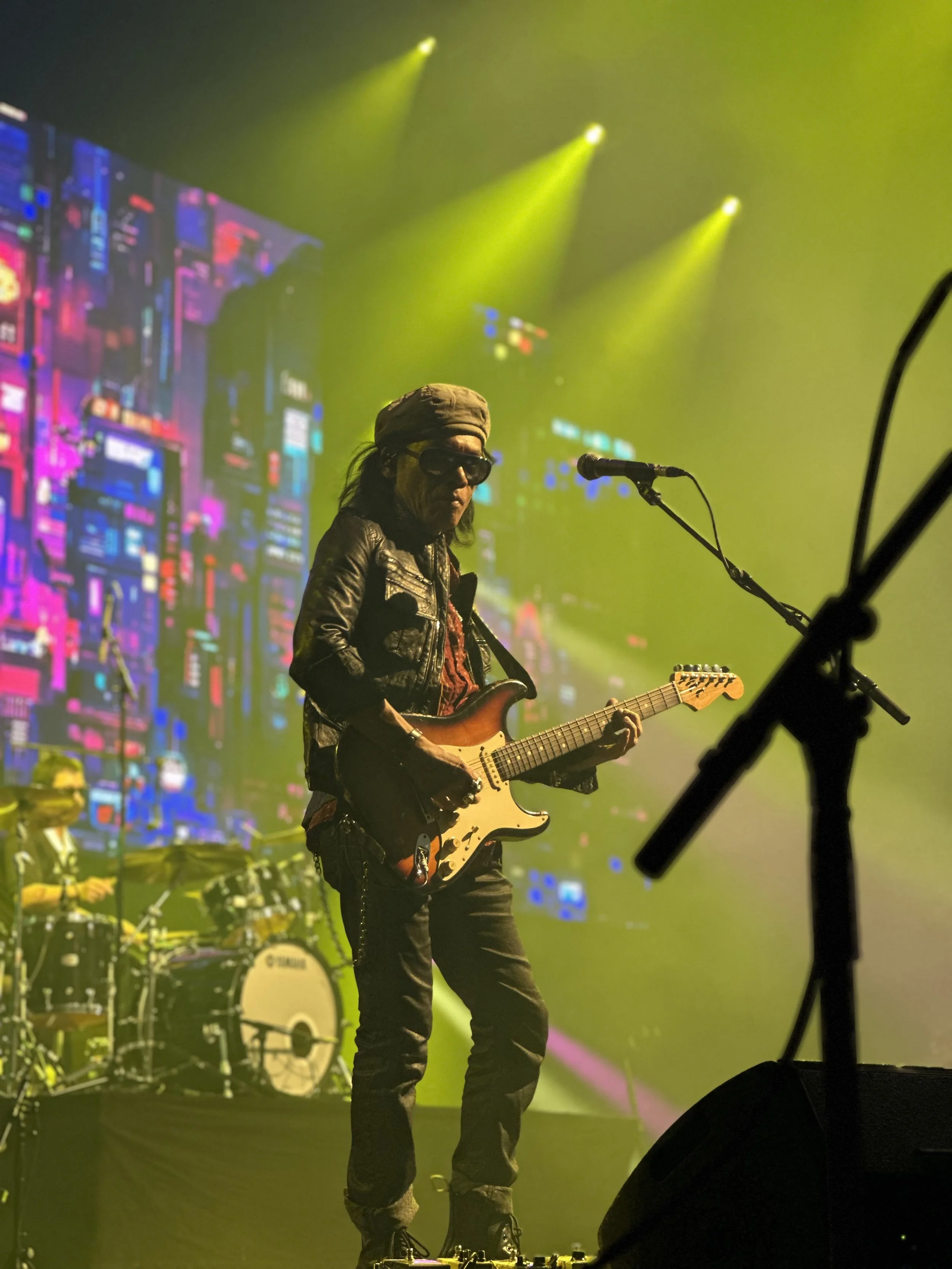 Musician playing electric guitar on stage with yellow lighting and a digital cityscape backdrop.