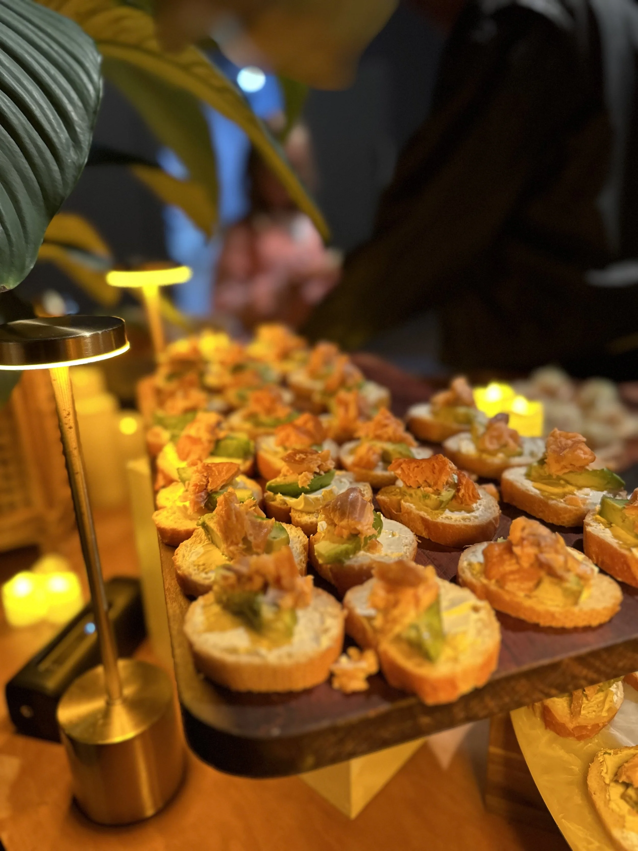 Assorted savory appetizers on a wooden tray, topped with green pickles and a creamy orange spread, under warm lighting.