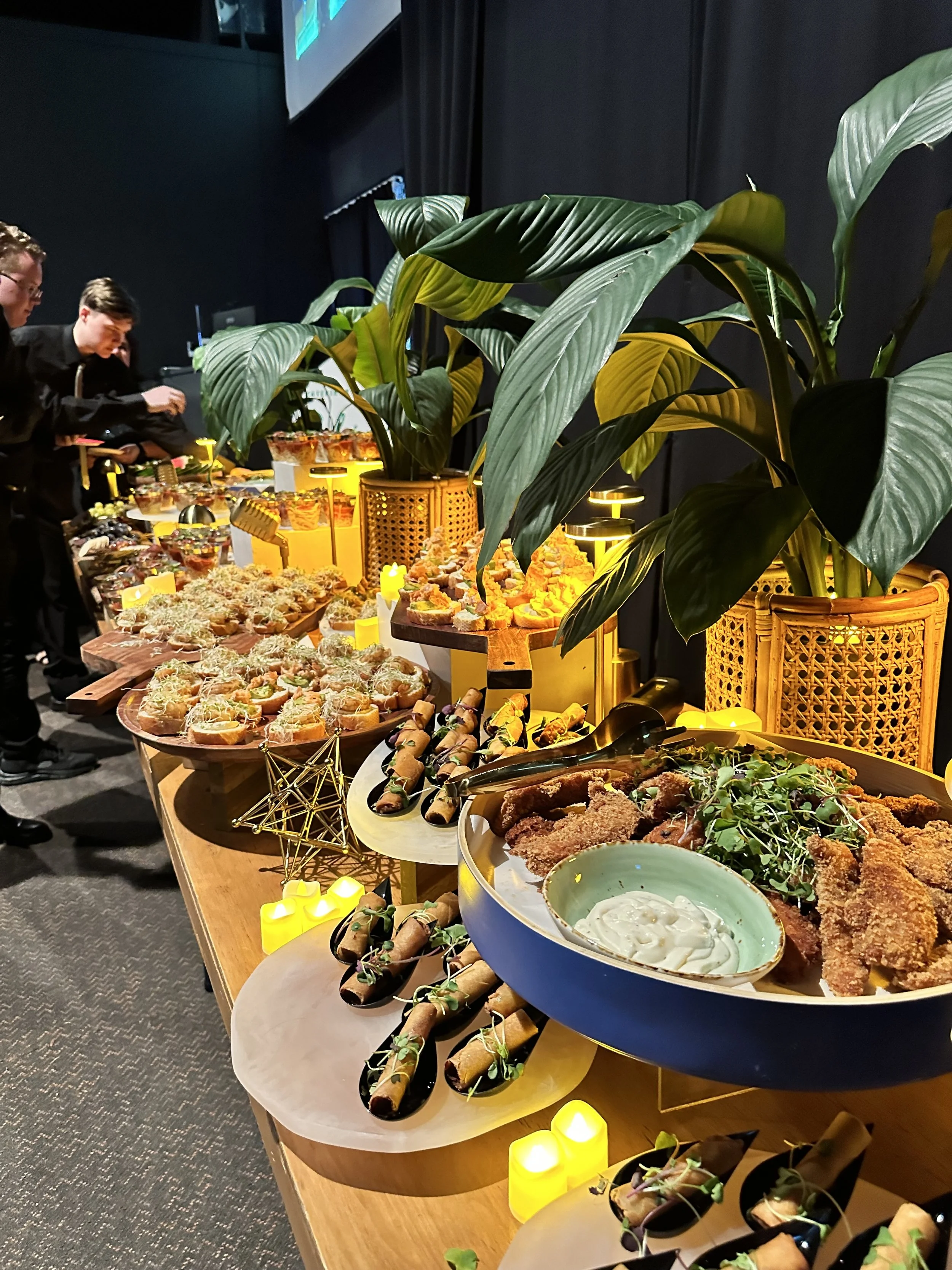 A buffet table with various appetizers and finger foods, including fried chicken, rolls, and garnished vegetables, decorated with yellow candles and large green plants in a dark room.