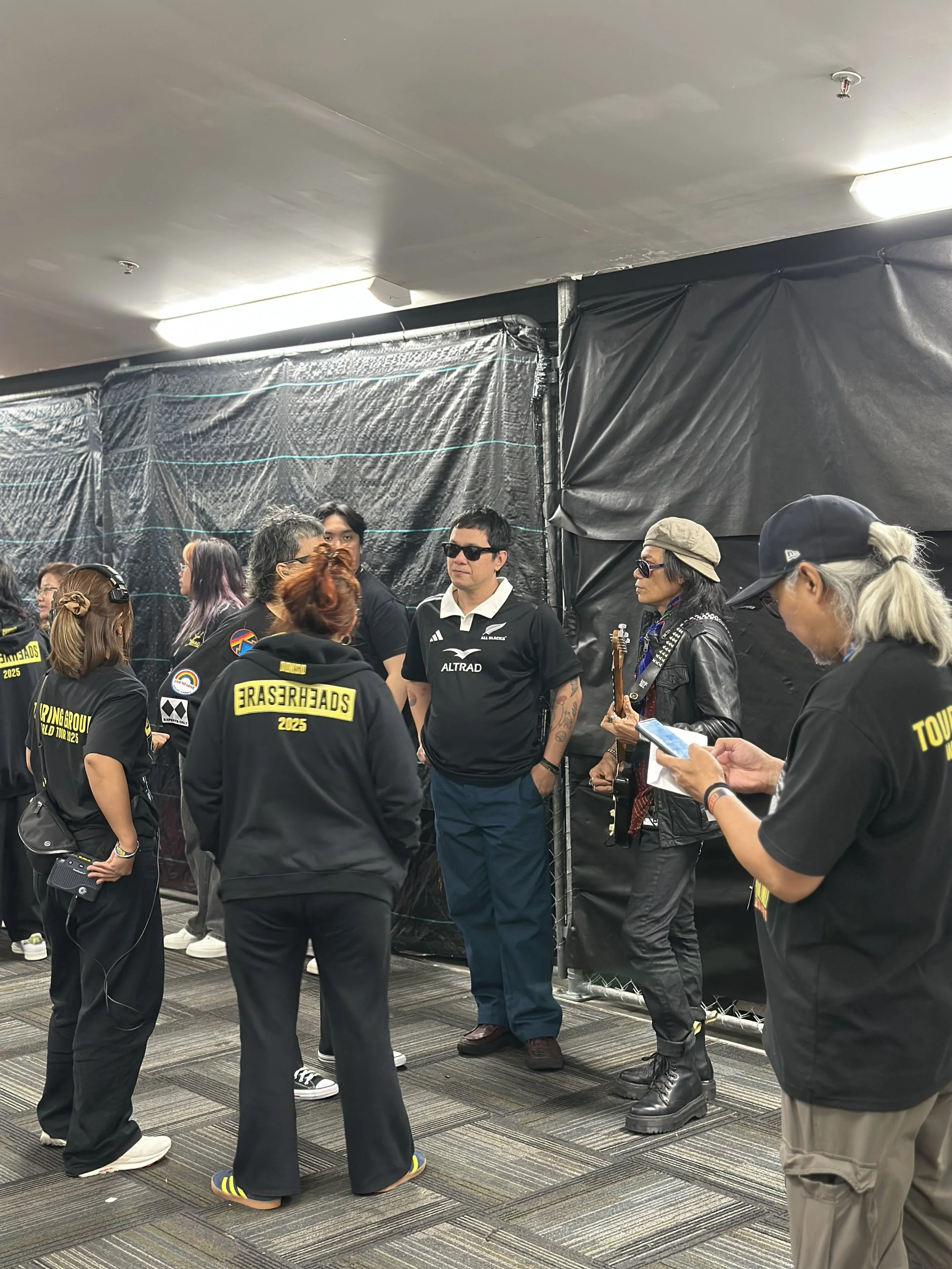 People gathered in a room, some wearing black jackets with 'TOURING GROUP 2025' printed on the back, others casually dressed. Some individuals are wearing sunglasses, and there is a woman holding a guitar. One person is looking at a smartphone. The b