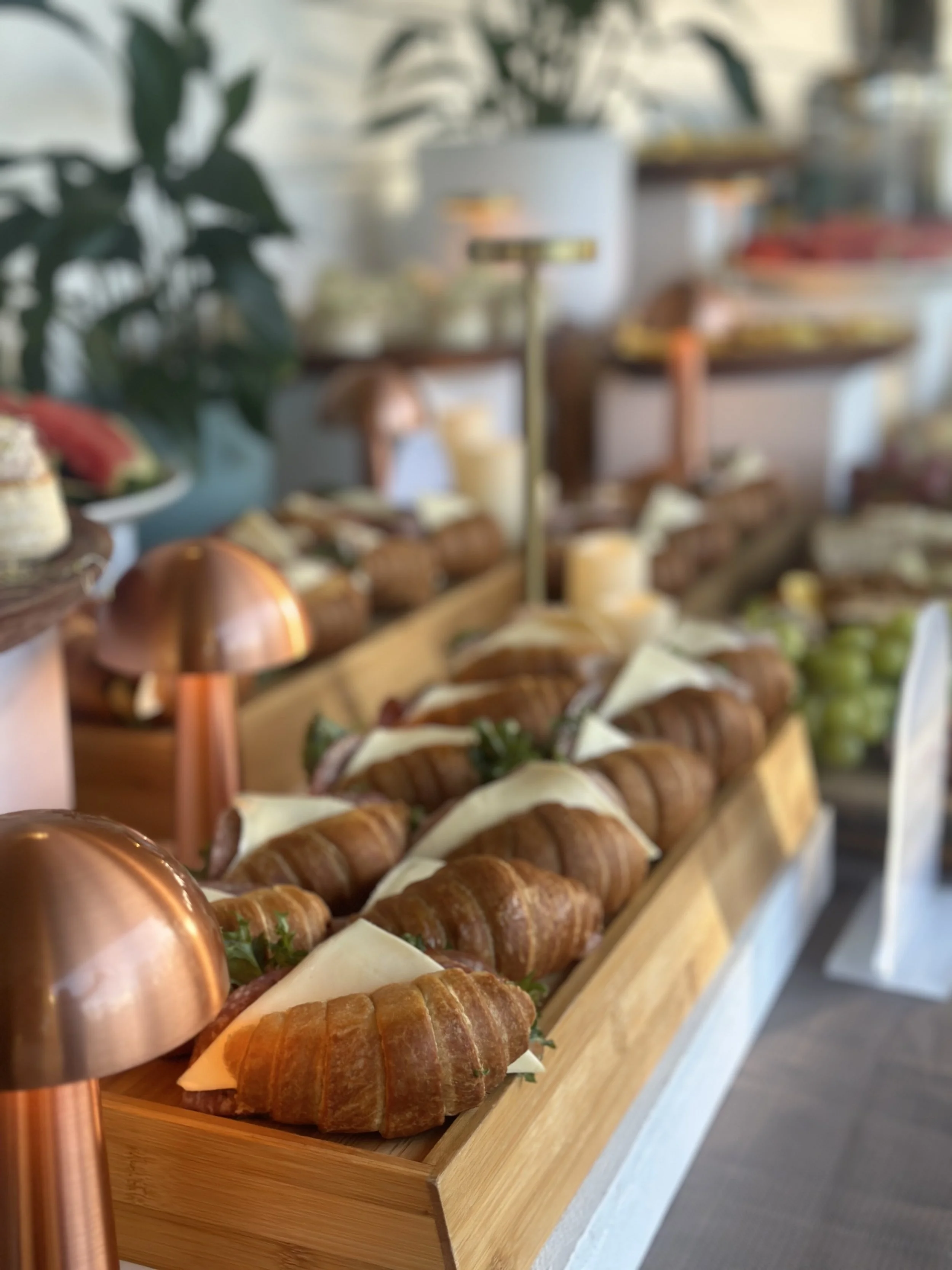 A display of croissant sandwiches with cheese and greens on a wooden tray, decorated with copper dome lamps in a cafe or bakery setting.