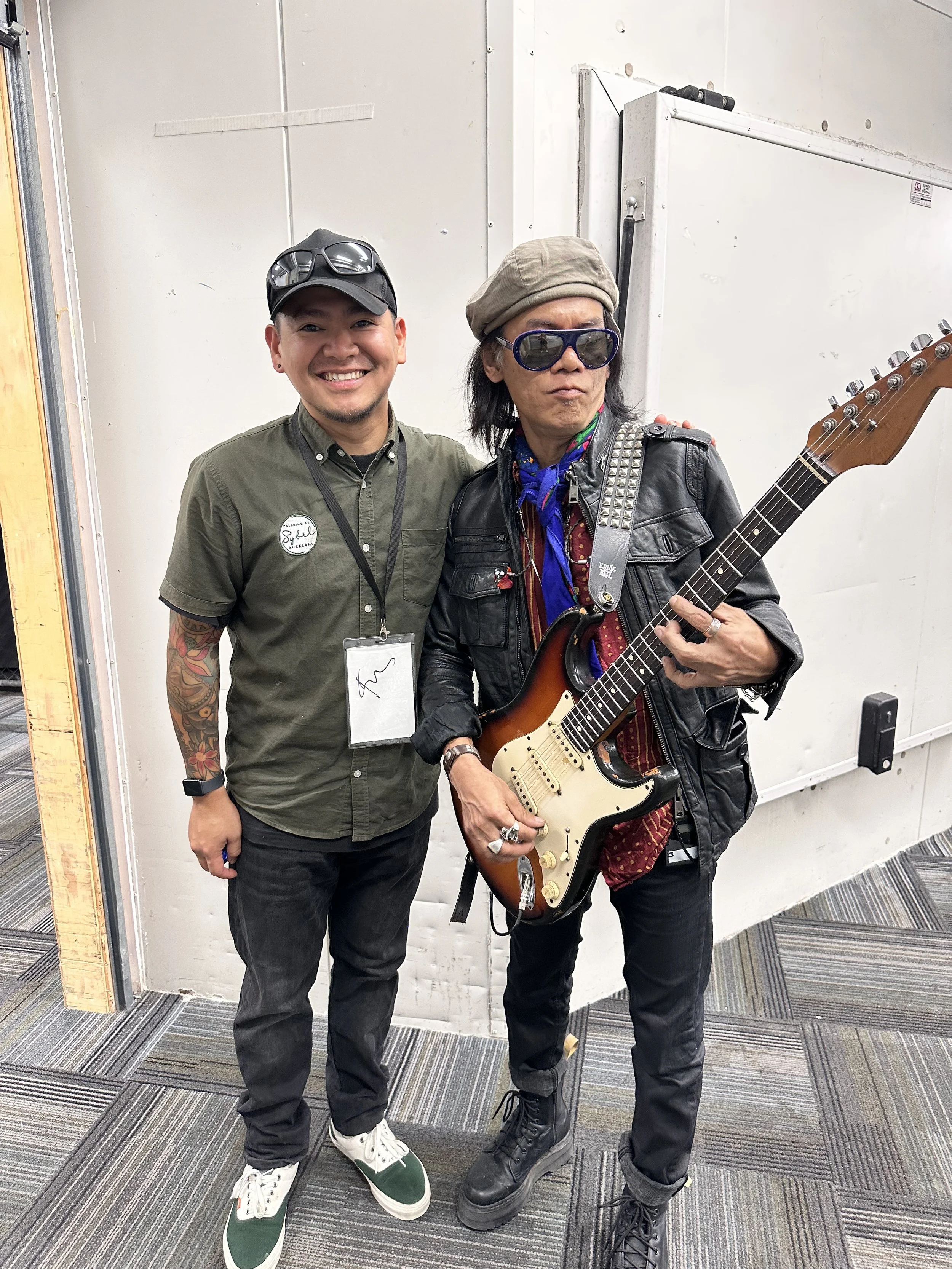 Two men standing together, one smiling and the other holding an electric guitar, in a room with white walls and carpeted floor.
