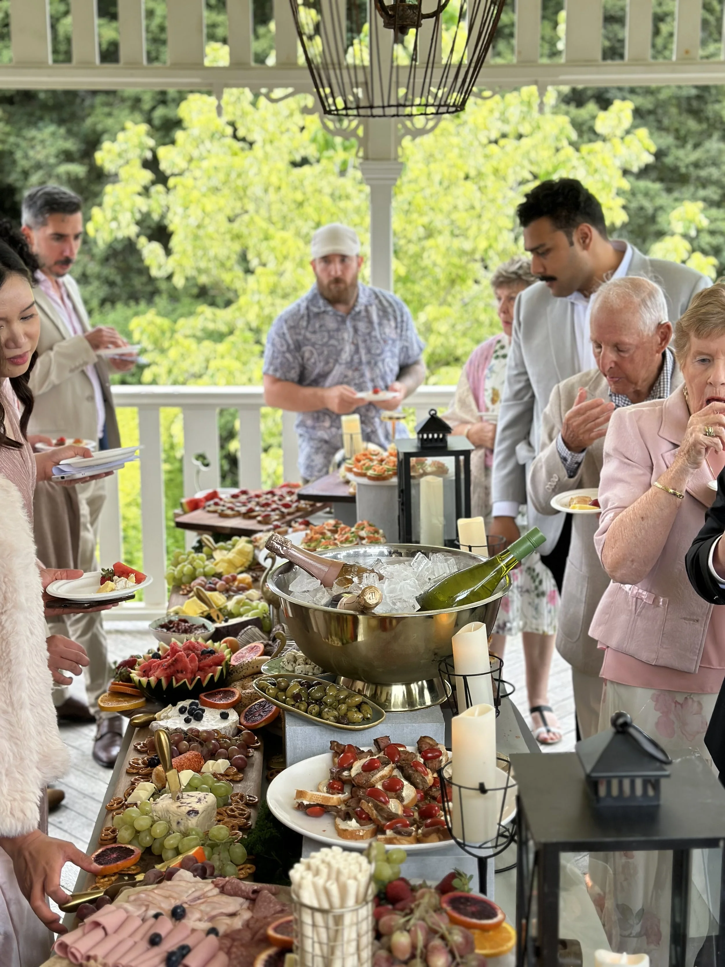 People gathering around a table filled with food and drinks at an outdoor event on a porch with green trees in the background.