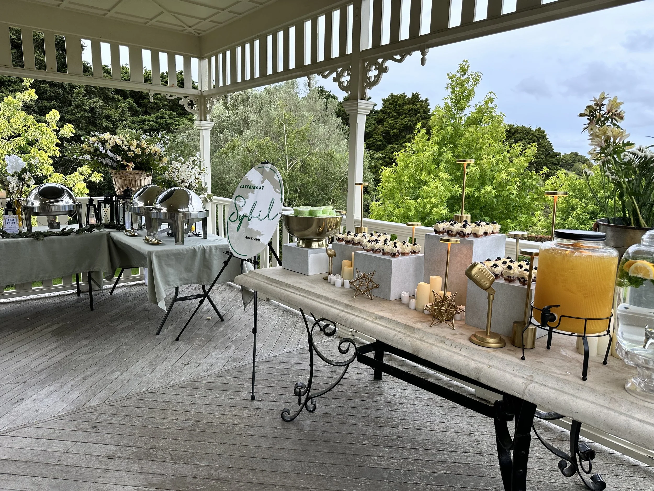 Buffet table setup on a covered porch with catering signs, chafing dishes, desserts, a lemonade dispenser, and floral arrangements, overlooking lush green trees.