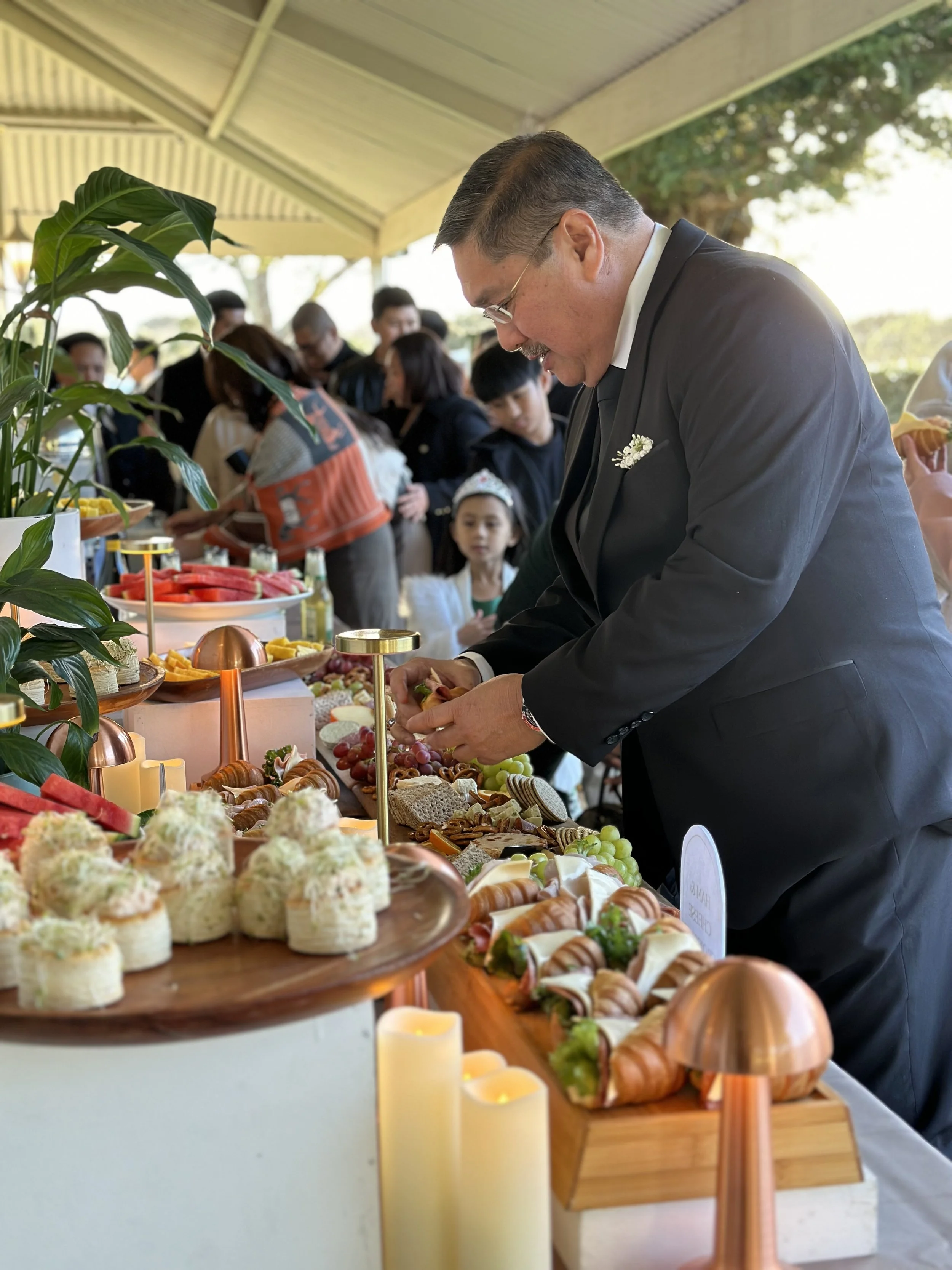 A man in a black suit selecting food from a buffet table at an outdoor event, with other guests in line behind him.
