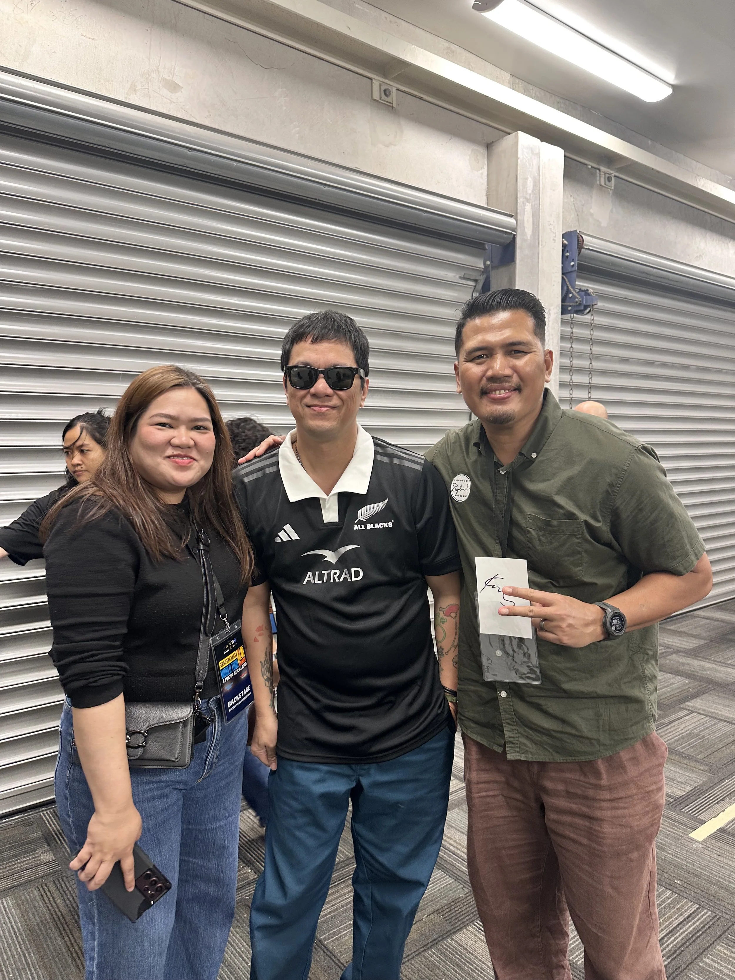 Three people standing together indoors in front of a roller shutter door, smiling at the camera. The person on the left is a woman with long brown hair, wearing a black jacket, blue jeans, and holding a phone. The person in the middle is a man wearin