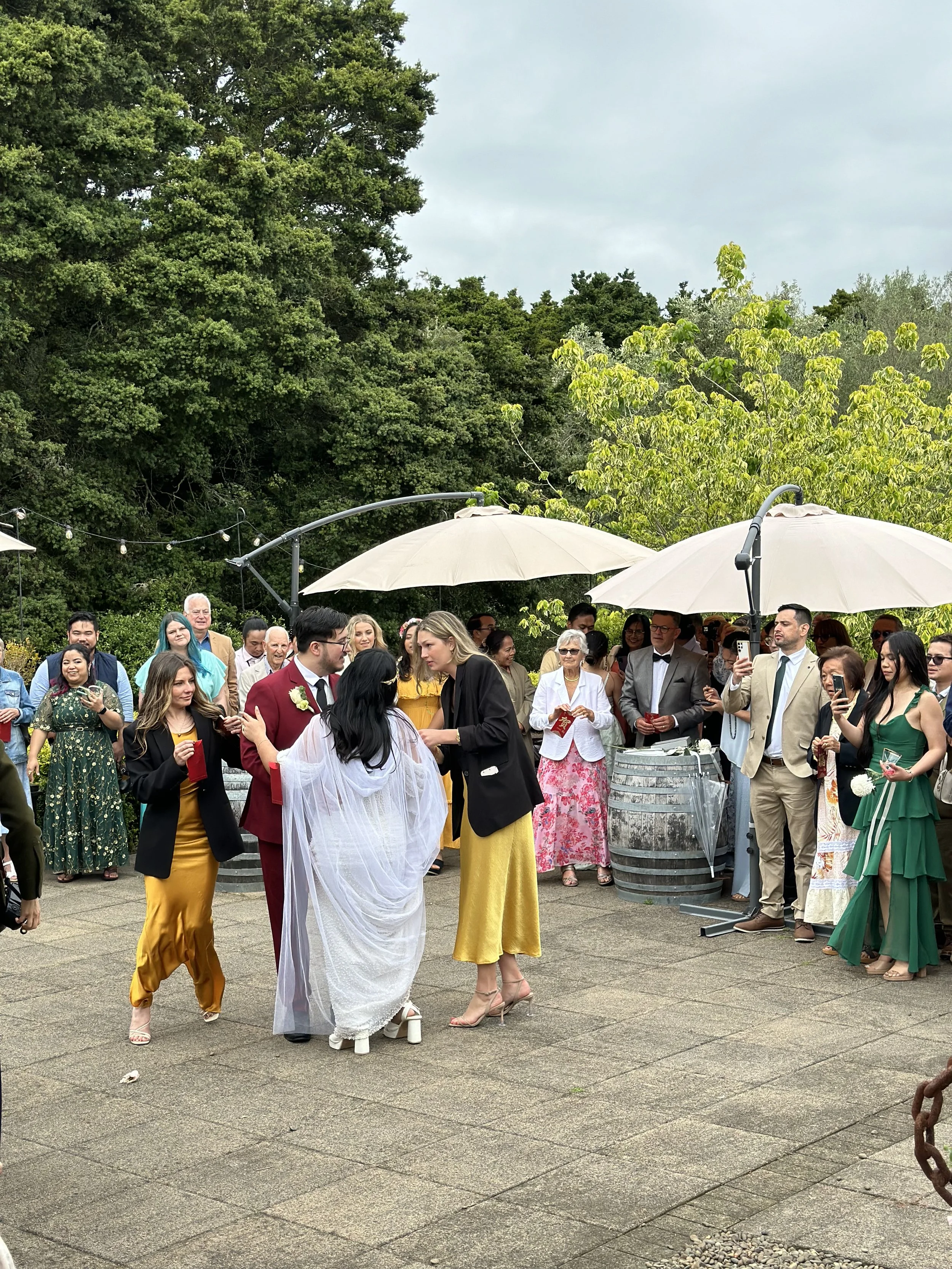 A wedding celebration outdoors with guests surrounding the bride and groom. The bride is in a white dress with a veil, and the groom is in a dark suit. Guests are holding drinks and taking photos, some under umbrellas. The setting is a garden area wi