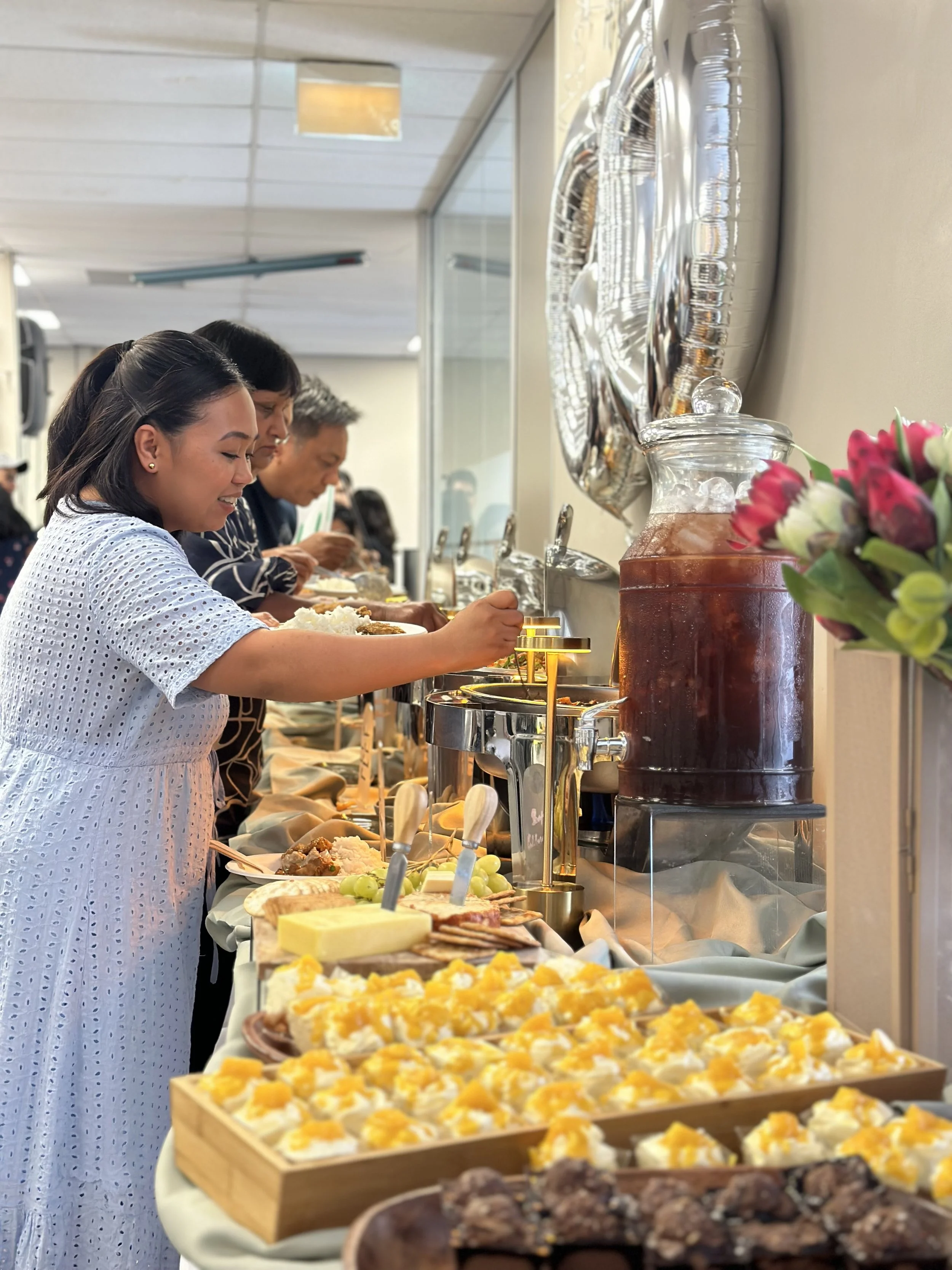 People serving themselves from a buffet with various dishes, including desserts topped with mango and cream, in a room with balloons and flowers.