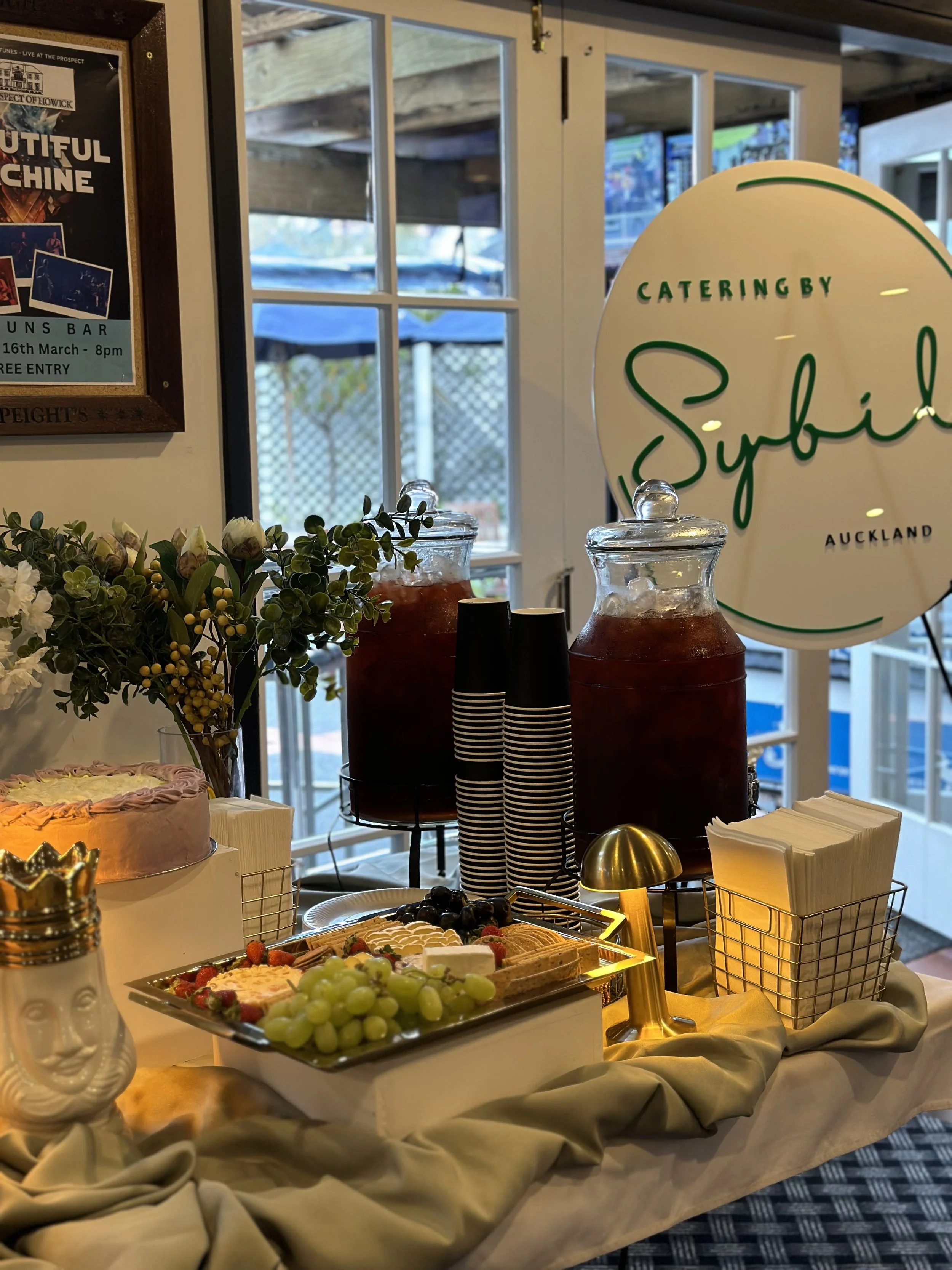 A buffet table with a floral arrangement, a cake, grapes, and assorted desserts, with two large jars of iced tea or juice, napkins in a wire holder, and a small golden lamp, set against a window with a sign reading 'Catering by Sybil' in green letter