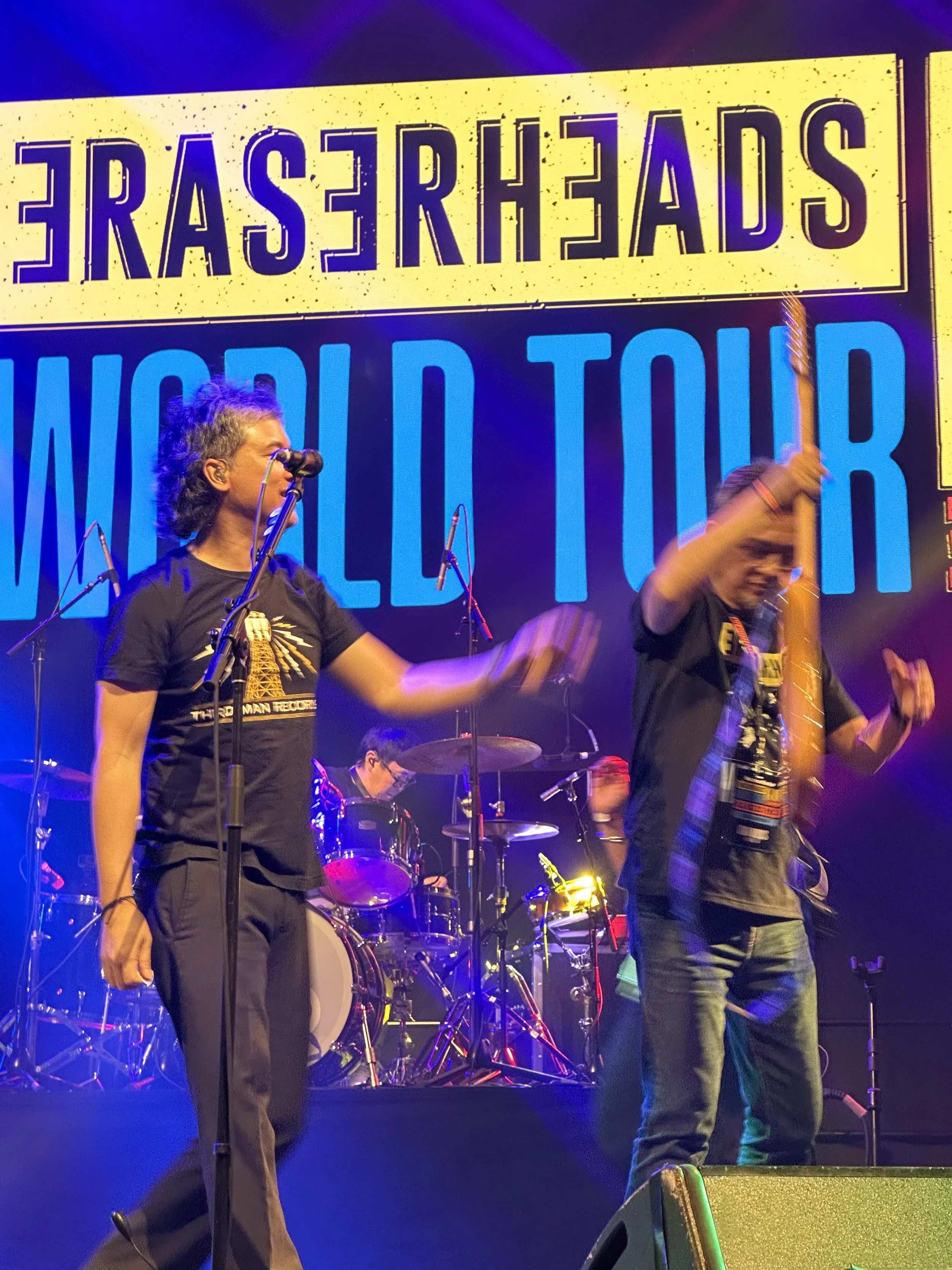 A live music performance on stage with a large screen behind reading 'ERASERHEADS WORLD TOUR'. The band members are actively engaging with instruments and microphone, with colorful stage lighting.