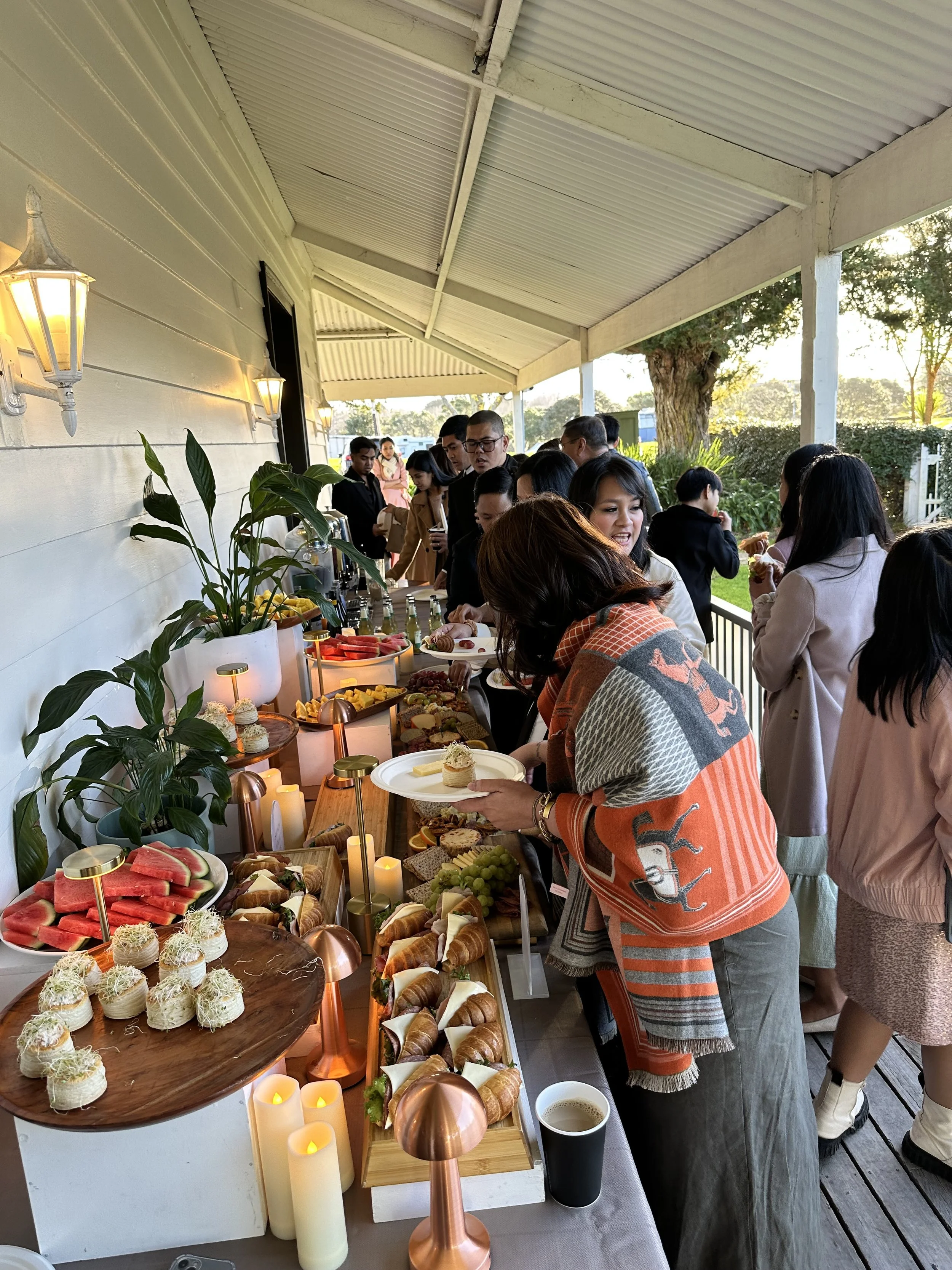 People at a buffet table outdoors enjoying food, with plates of pastries, fruits, and desserts, decorated with plants and candles.
