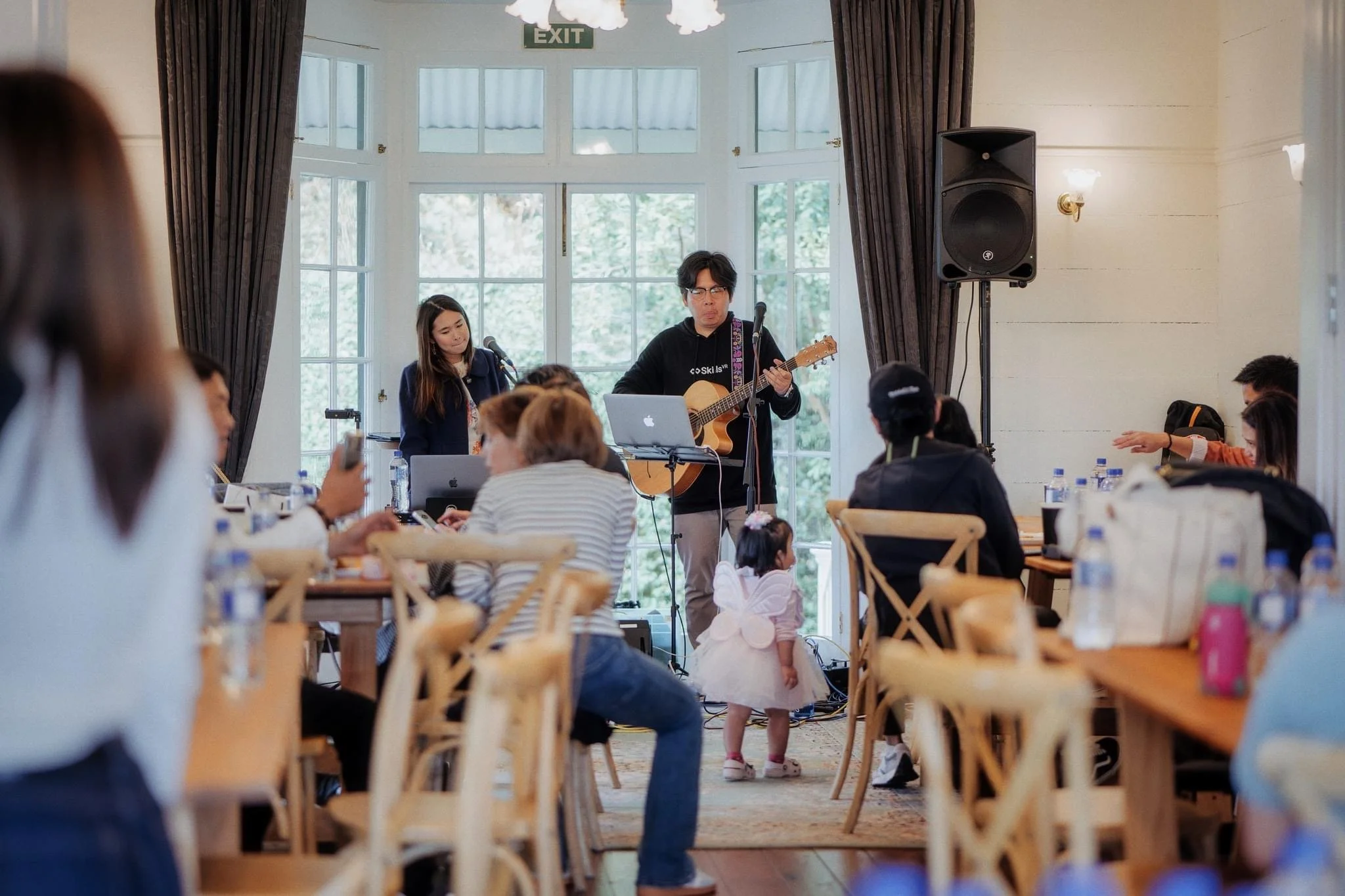 A man playing guitar and singing into a microphone with a woman beside him at a live music performance in a well-lit room with large windows and curtains, surrounded by seated audience members and a small child in a tutu watching the performance.